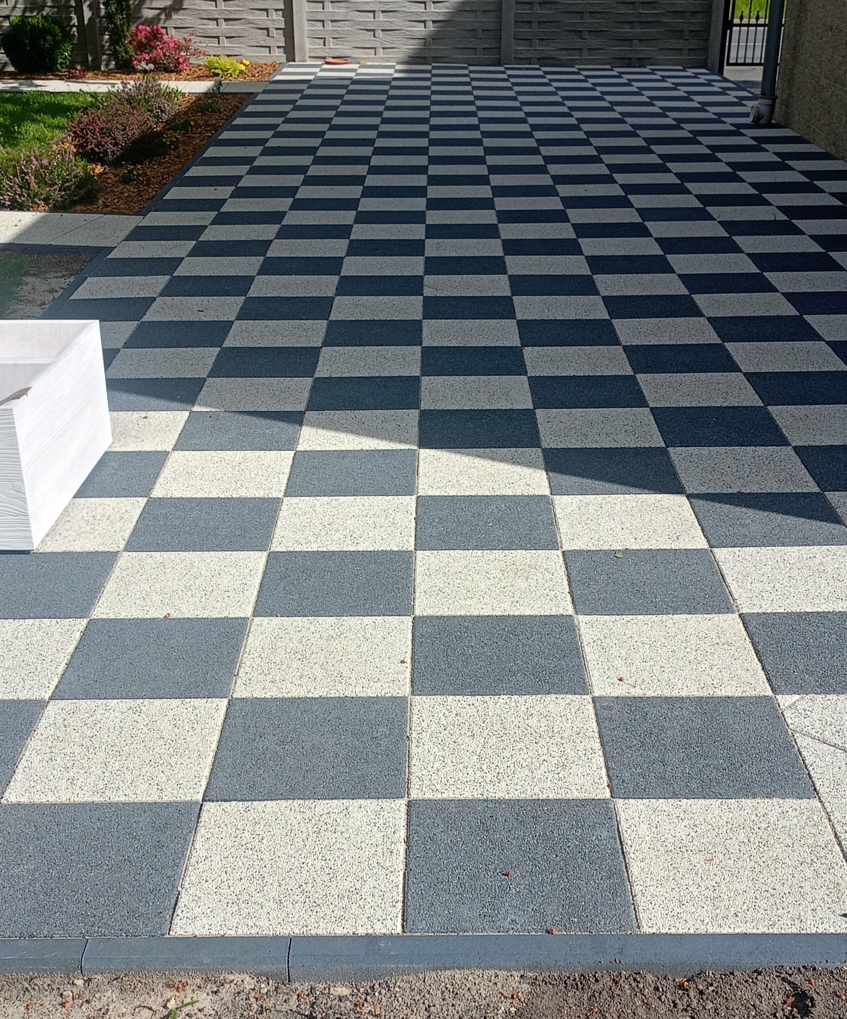 Szachownica z kostki brukowej w odcieniach szarości i bieli, prowadząca do bramy. Widoczne obrzeża i fragment rabaty z roślinami. Ujęcie z niskiej perspektywy.
