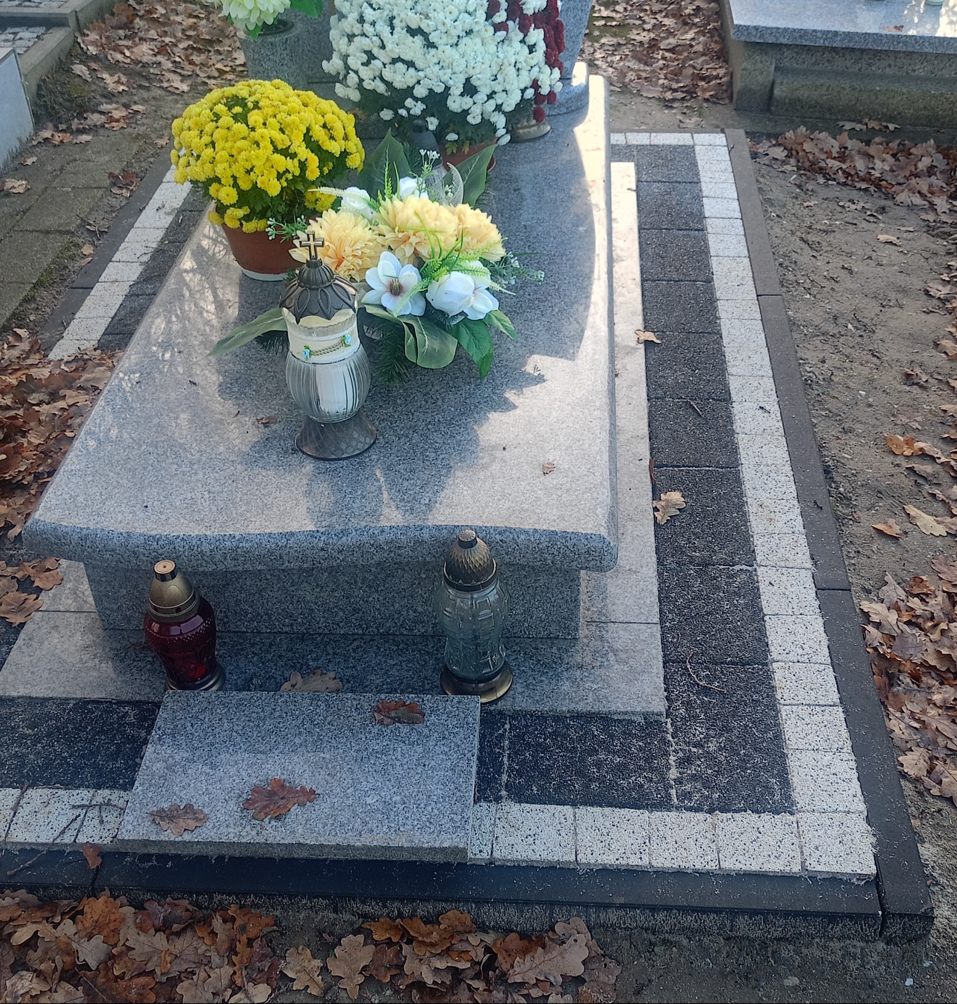 Granitowy nagrobek z obrzeżem z kostki brukowej w odcieniach szarości, ozdobiony kwiatami i zniczami, otoczony opadłymi liśćmi.