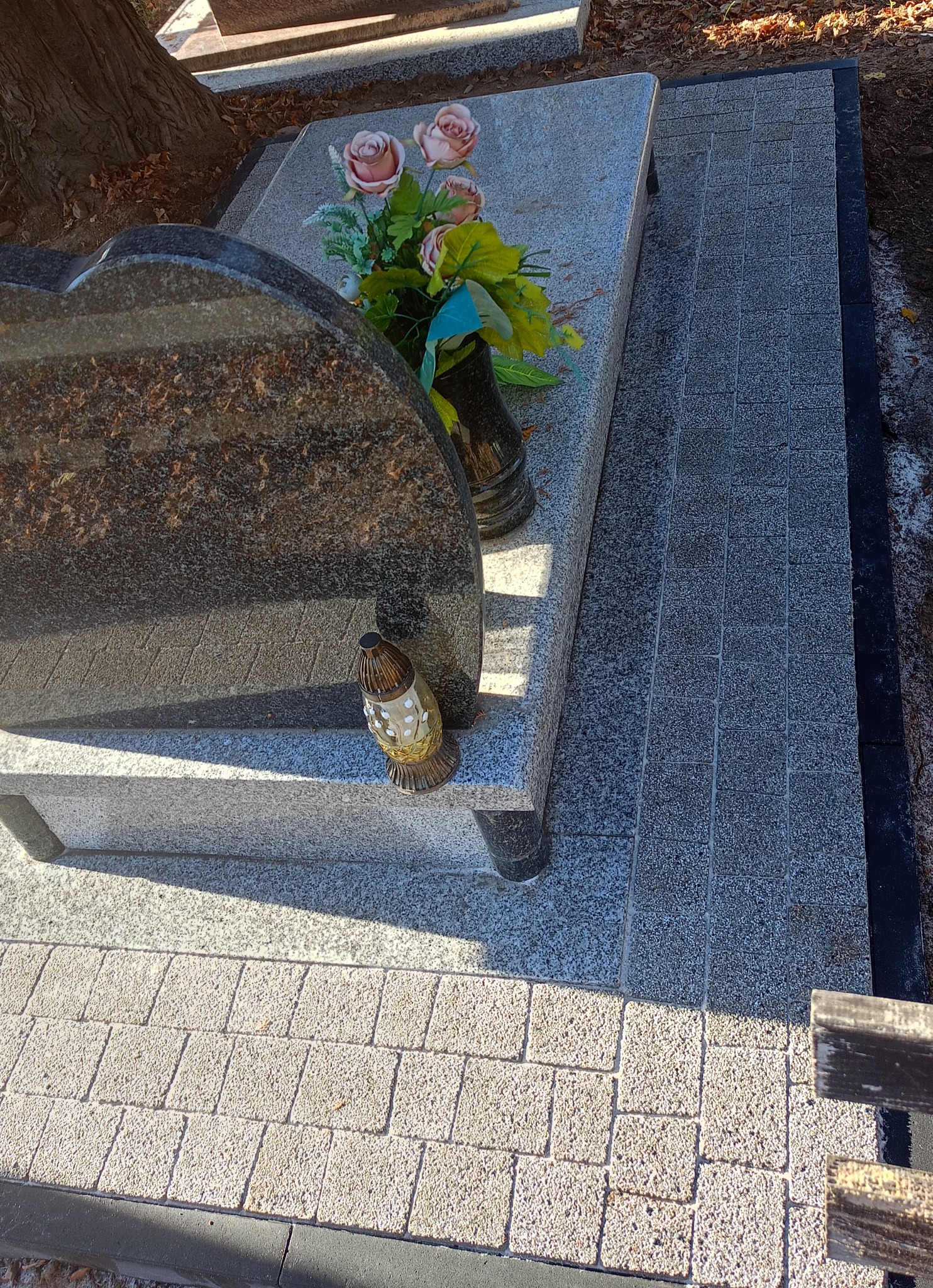 Granitowy nagrobek z kwiatami i lampionem, otoczony jasnoszarą kostką brukową. Ciemnoszara obwódka kostki. Ujęcie z góry, widoczny fragment drzewa.