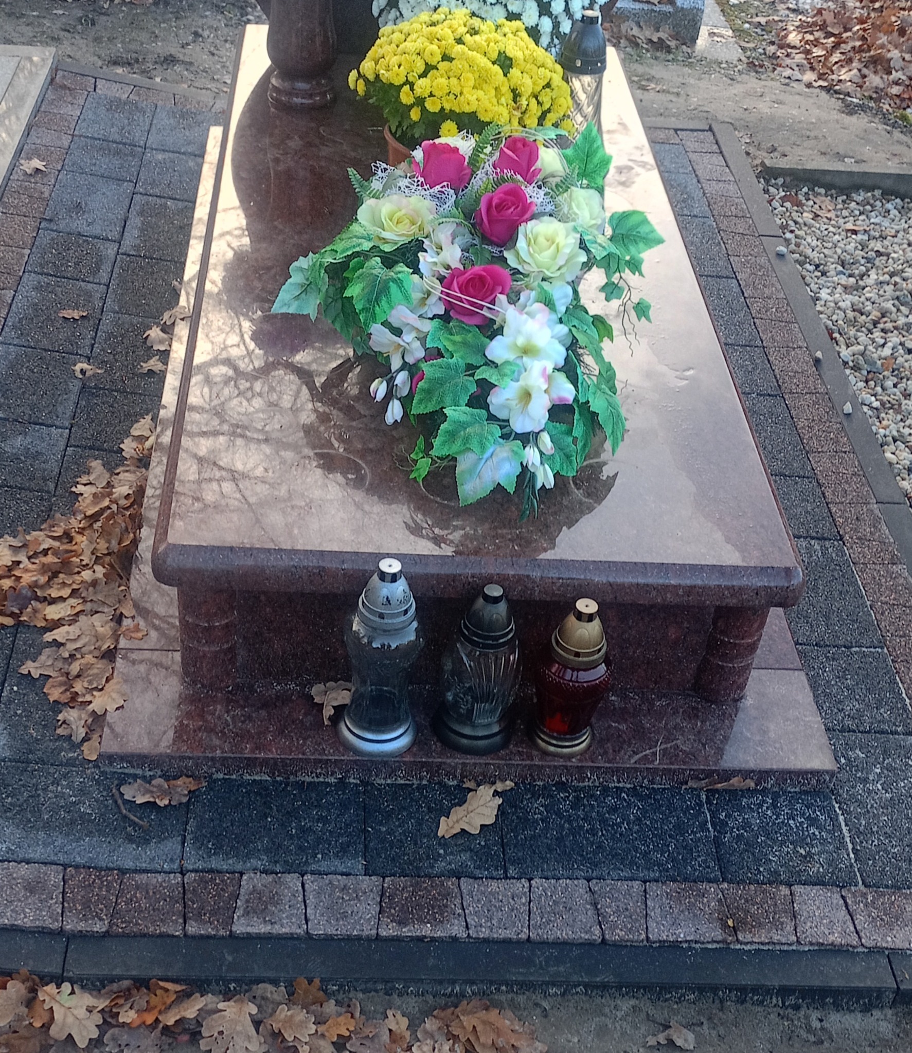 Granitowy nagrobek z kwiatami i zniczami, otoczony kostką brukową w jesiennej scenerii. Widoczne liście na kostce brukowej.