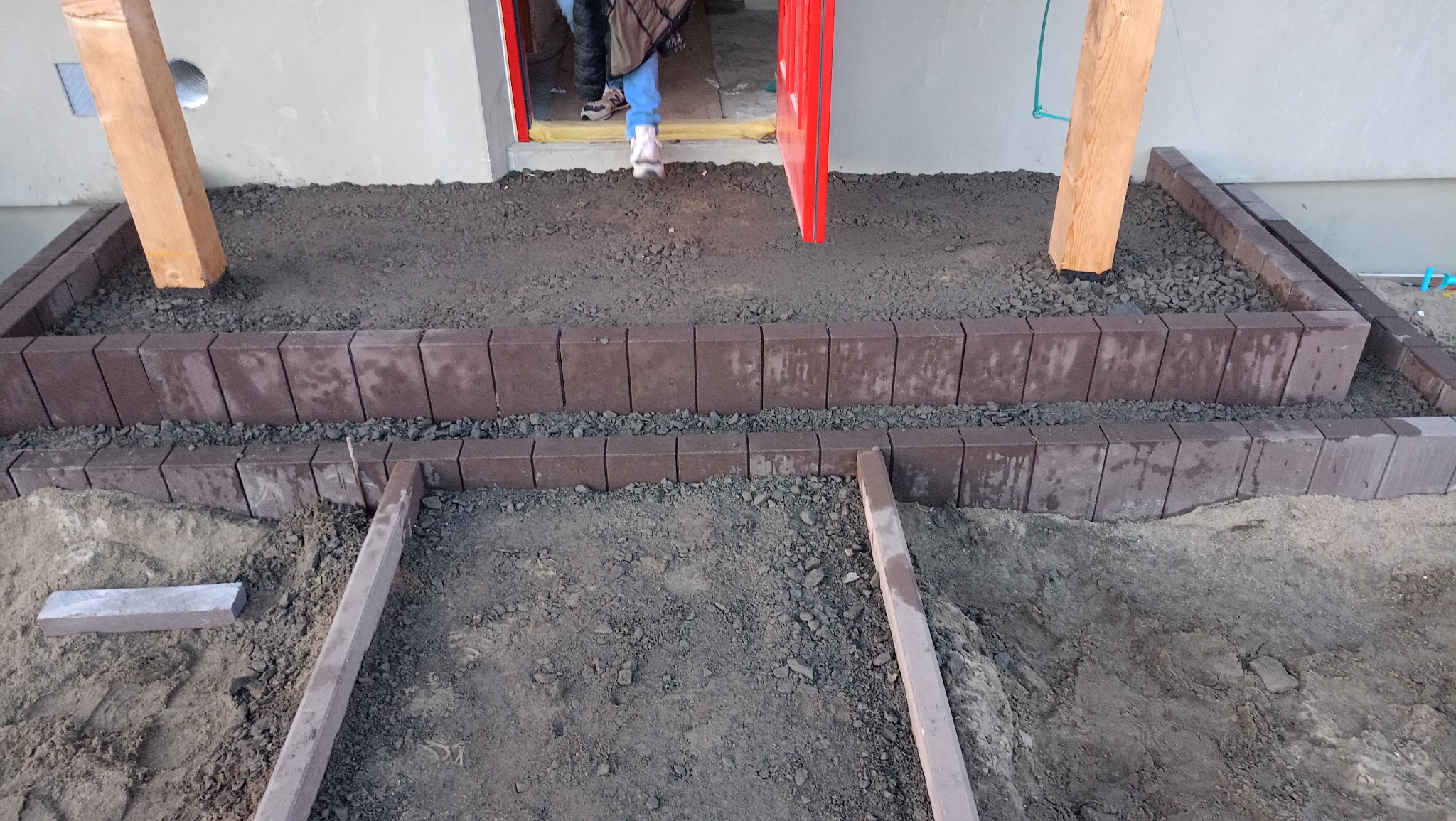 Układanie brązowej kostki brukowej wokół drewnianych słupów, widoczne etapy prac przygotowawczych przed wejściem do budynku z czerwoną ramą drzwi.