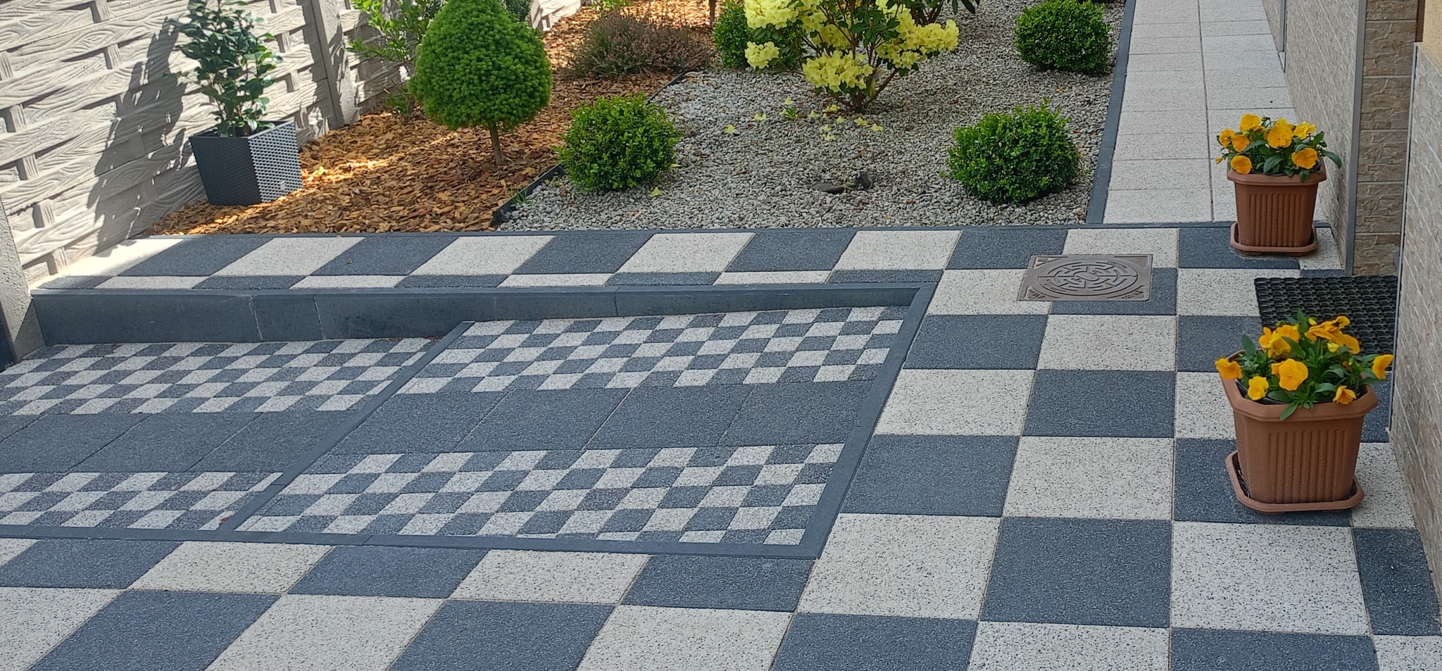 Szachownica z kostki brukowej w odcieniach szarości i beżu, z obrzeżami i schodkiem. W tle roślinność i donice z żółtymi kwiatami. Widok z góry.