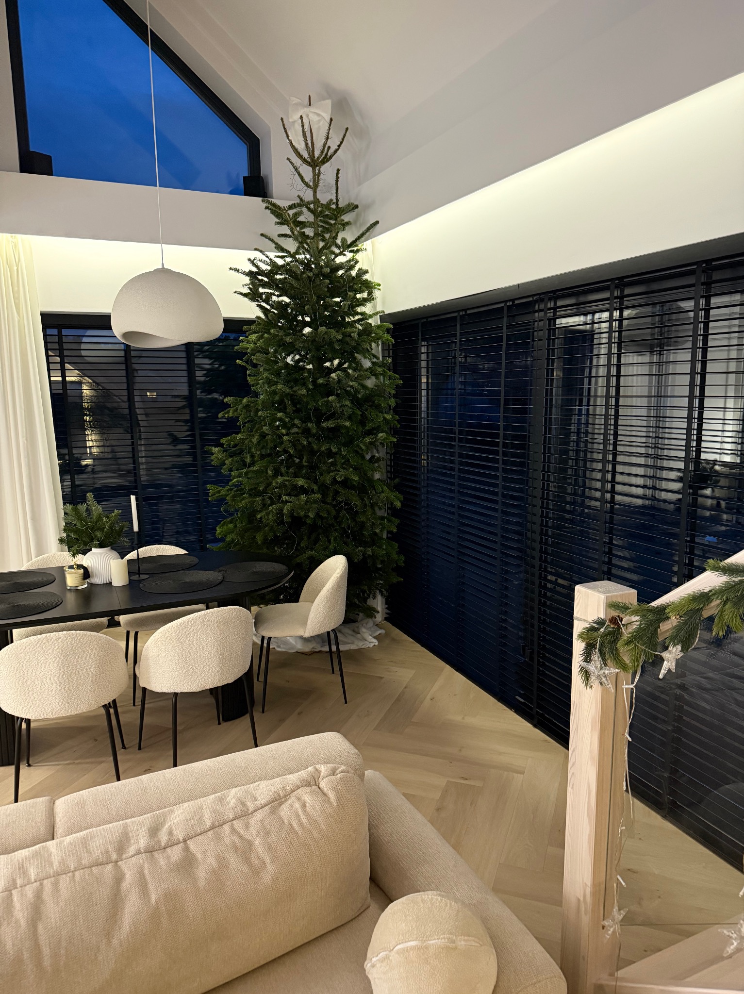 Nowoczesne, czarne żaluzje aluminiowe w salonie z choinką, widoczne z perspektywy sofy. Minimalistyczny design, ciepłe oświetlenie, parkiet w jodełkę.