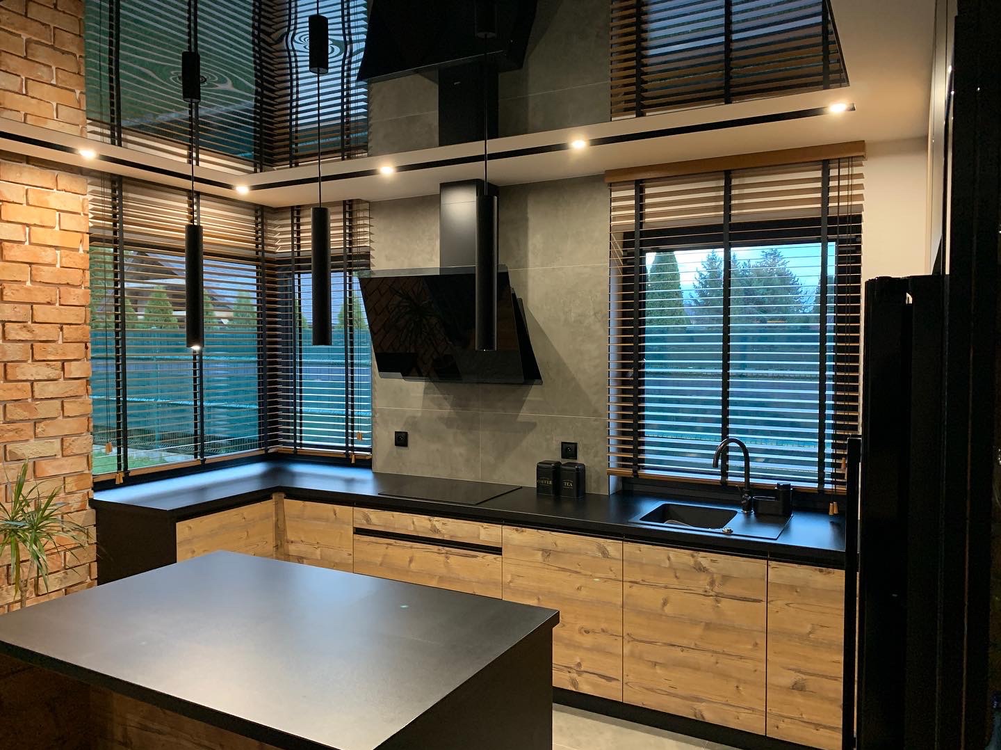 Nowoczesna kuchnia z drewnianymi żaluzjami poziomymi w oknach, czarnym blatem i okapem. Widoczna ceglana ściana i wyspa kuchenna.