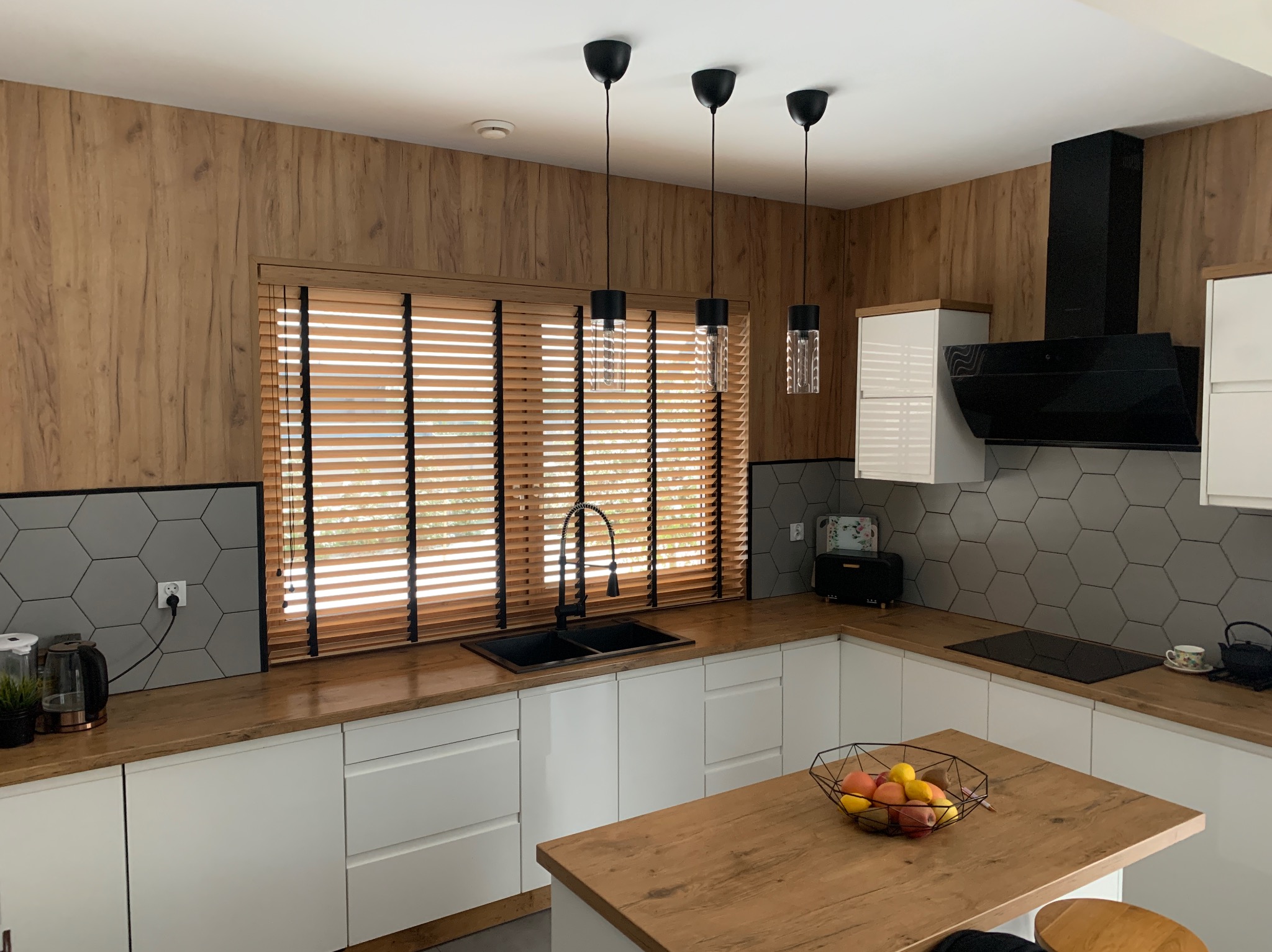 Kuchnia z drewnianymi żaluzjami poziomymi w oknie, czarną armaturą i heksagonalnymi płytkami. Widoczne trzy lampy wiszące i drewniane blaty.
