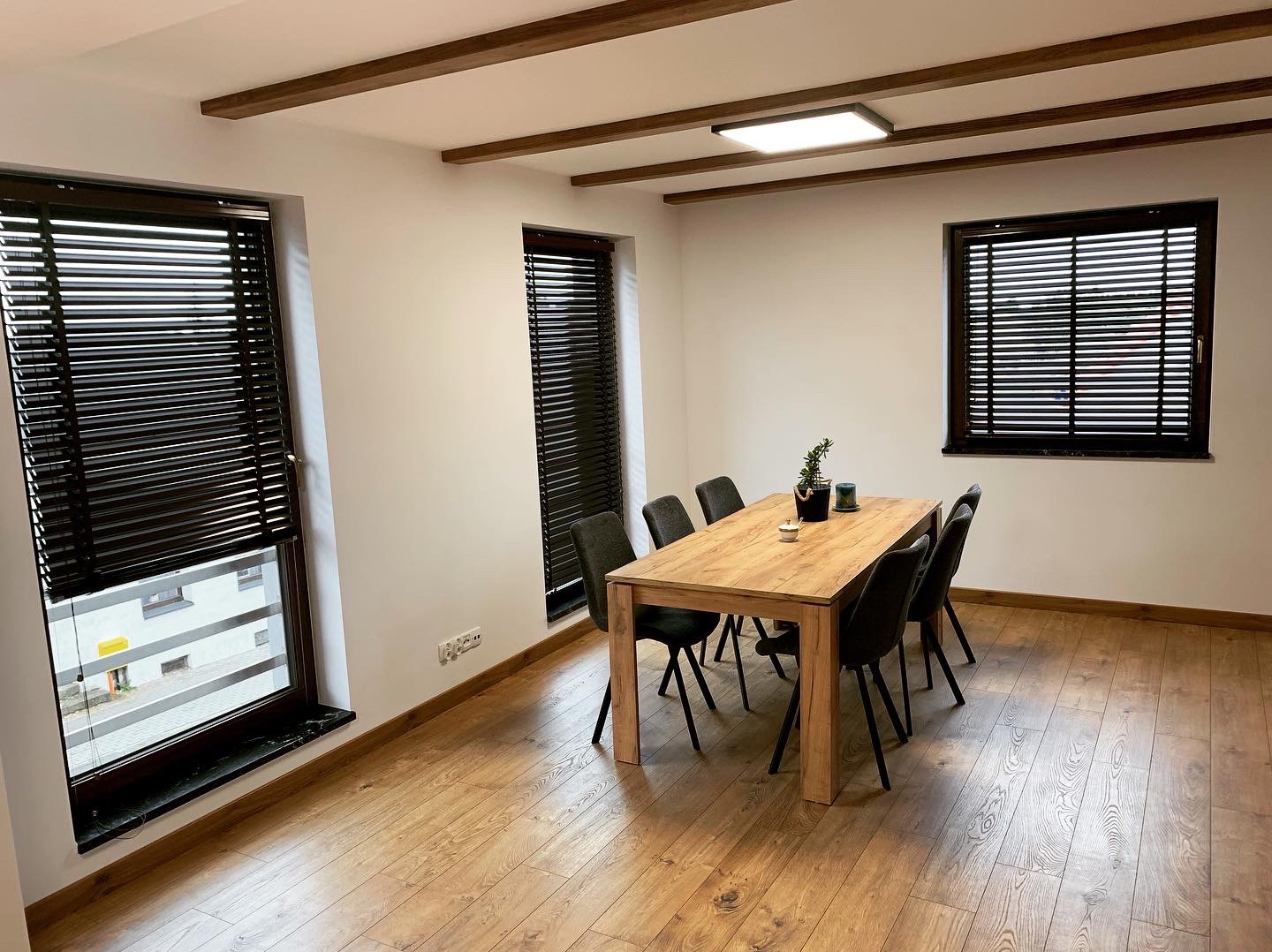 Jadalnia z trzema oknami z ciemnymi żaluzjami drewnianymi. Widok na zewnątrz z jednego okna. Drewniany stół i krzesła. Drewniane belki na suficie.