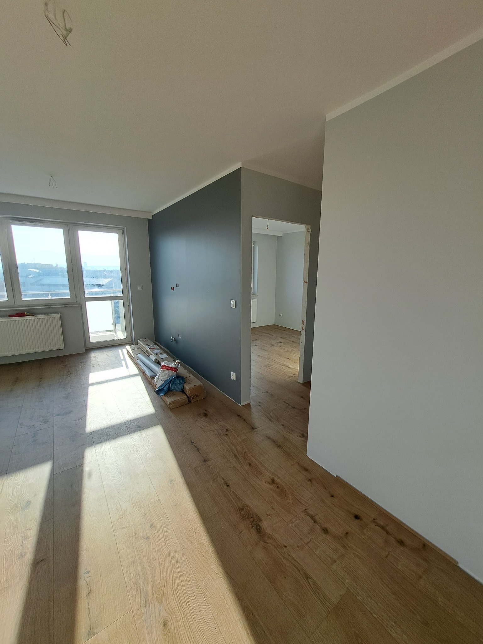 Jasne, przestronne wnętrze po remoncie: drewniana podłoga, szara ściana akcentująca, okno balkonowe z widokiem na miasto. Przy ścianie materiały wykończeniowe.