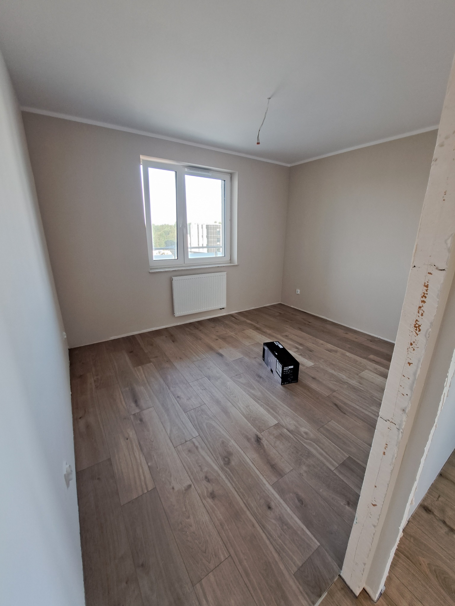 Jasne wnętrze po remoncie: brązowe panele podłogowe, beżowe ściany, białe okno z grzejnikiem. Widoczna skrzynka na podłodze i fragment ościeżnicy.