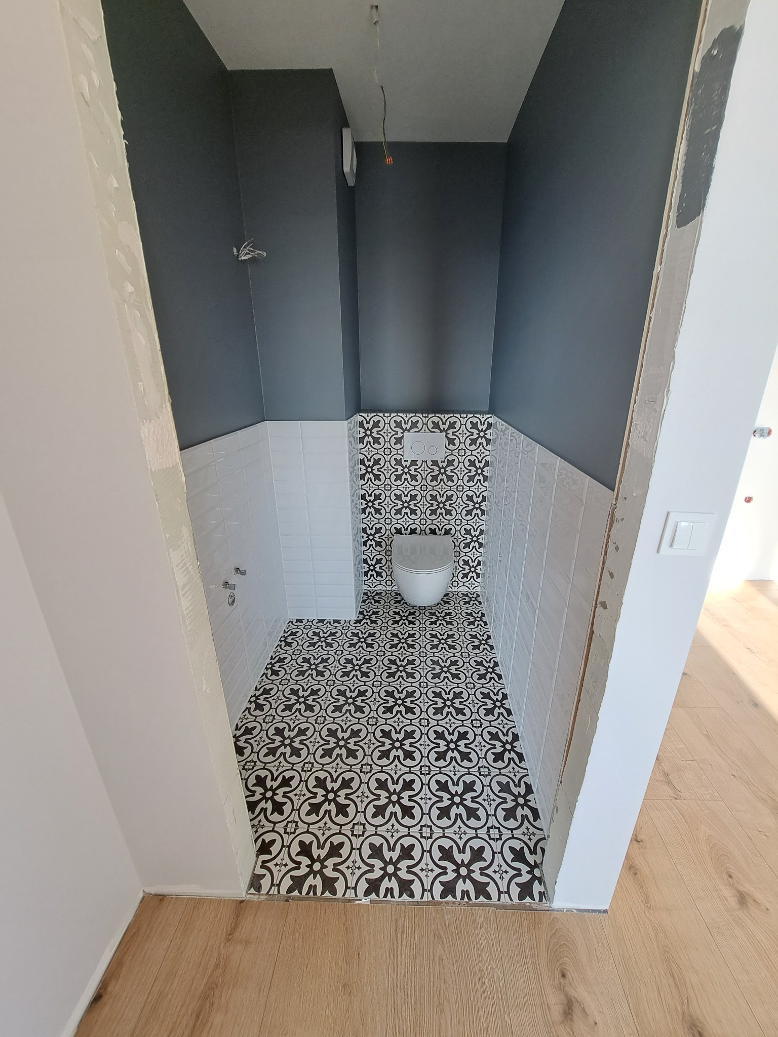 Wąska toaleta po remoncie: czarno-biała mozaika na podłodze i ścianie za toaletą, białe kafelki i szara farba na ścianach. Widok z korytarza na drewnianą podłogę.