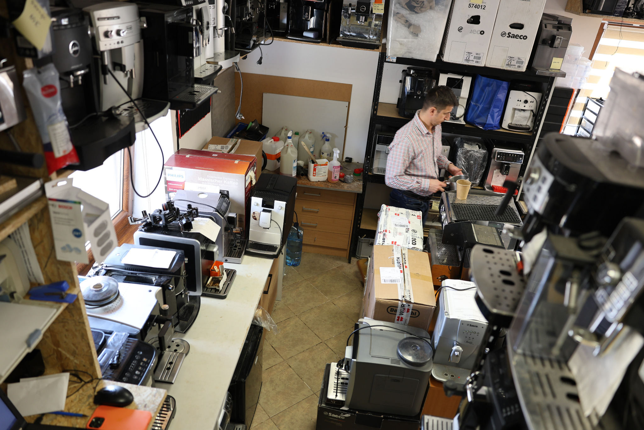 Serwis ekspresów: inżynier Grzegorz z IventSerwis.pl w warsztacie, przygotowuje kawę. W tle półki z ekspresami i częściami zamiennymi, widok z góry.