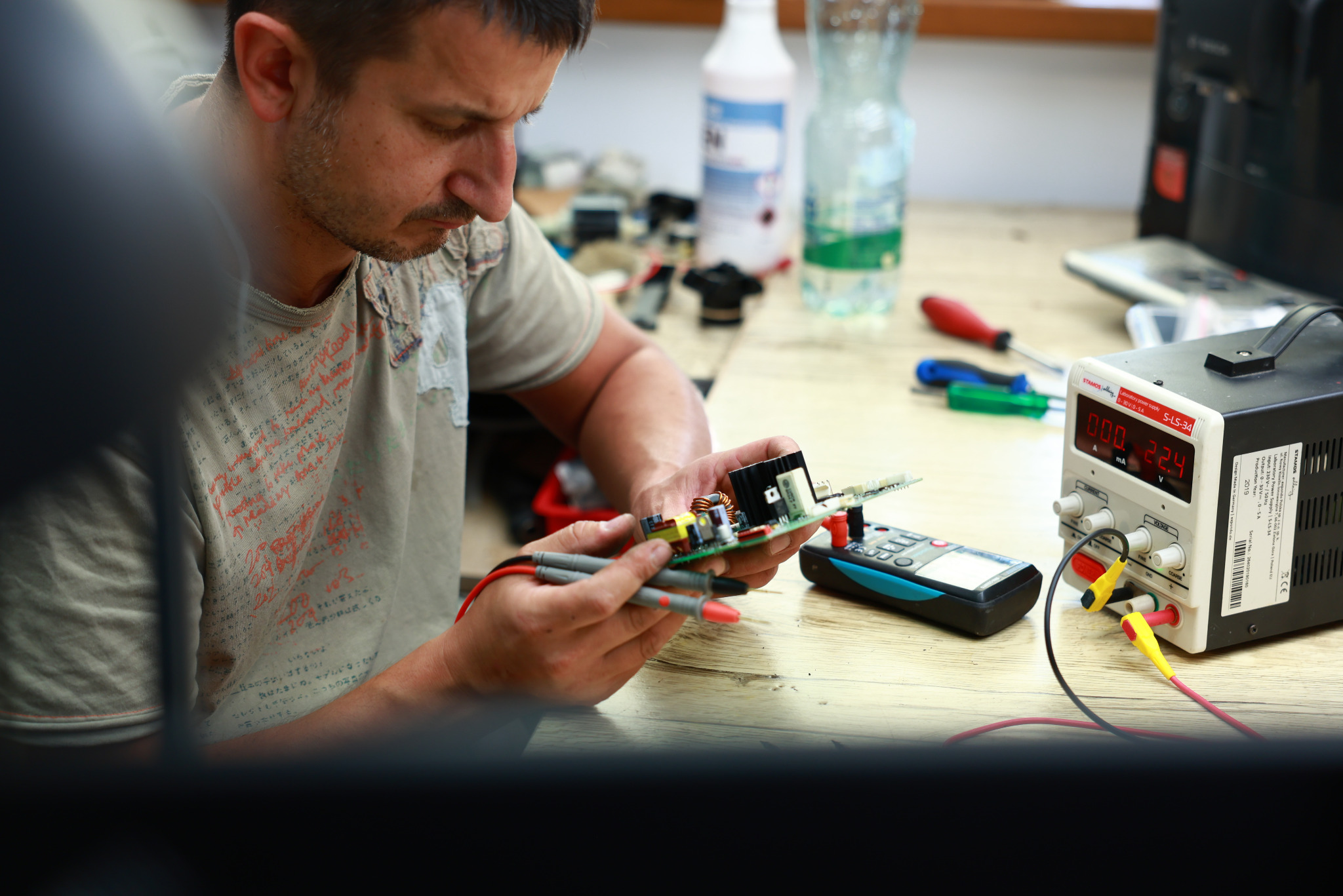 Serwisant diagnozuje płytę główną ekspresu Saeco Lirika za pomocą multimetru i zasilacza laboratoryjnego. Widoczne narzędzia i elementy elektroniczne na stole.