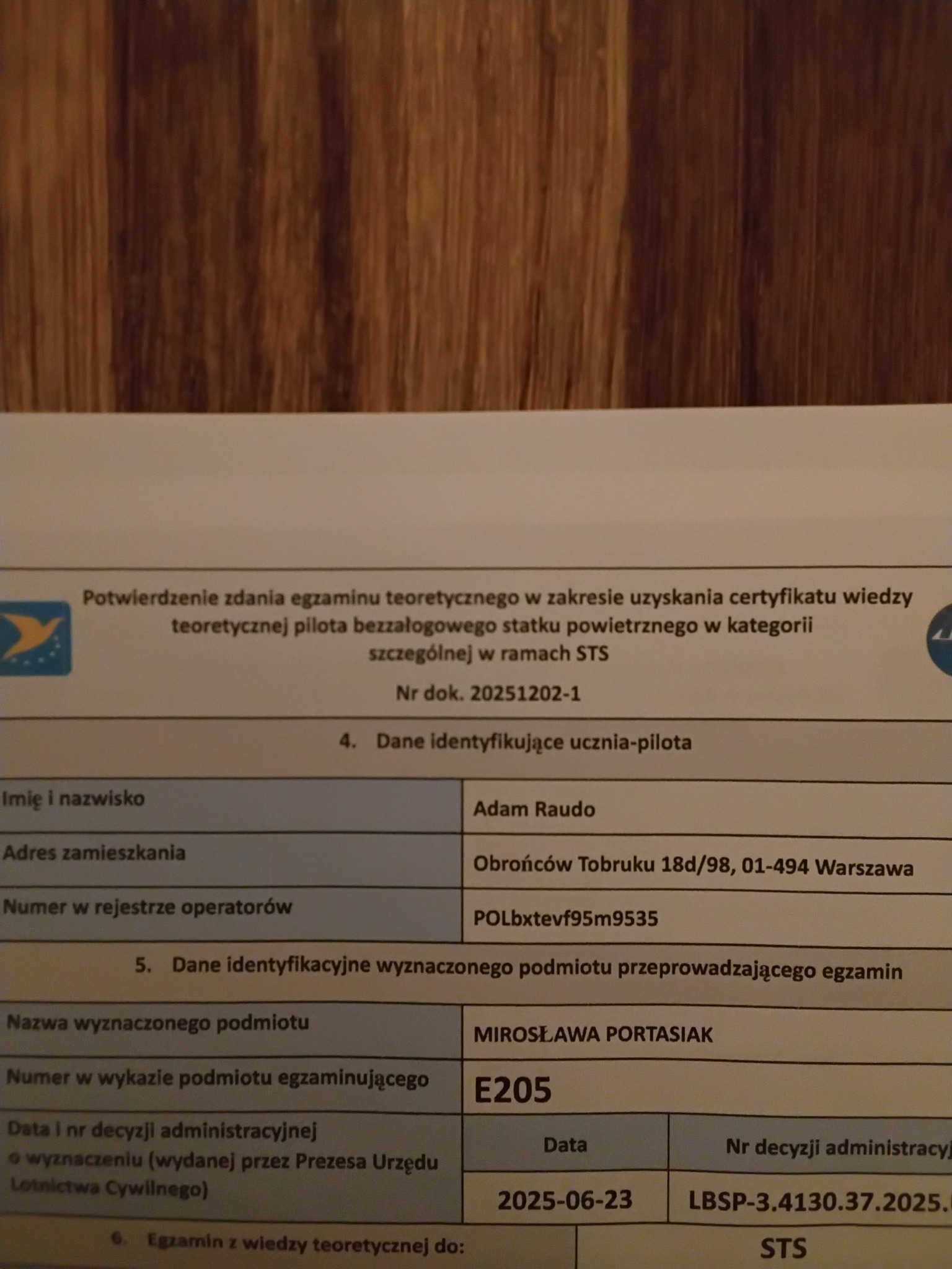 Dokument potwierdzający zdanie egzaminu teoretycznego na pilota drona, z danymi osobowymi i numerem dokumentu, leżący na drewnianej powierzchni.