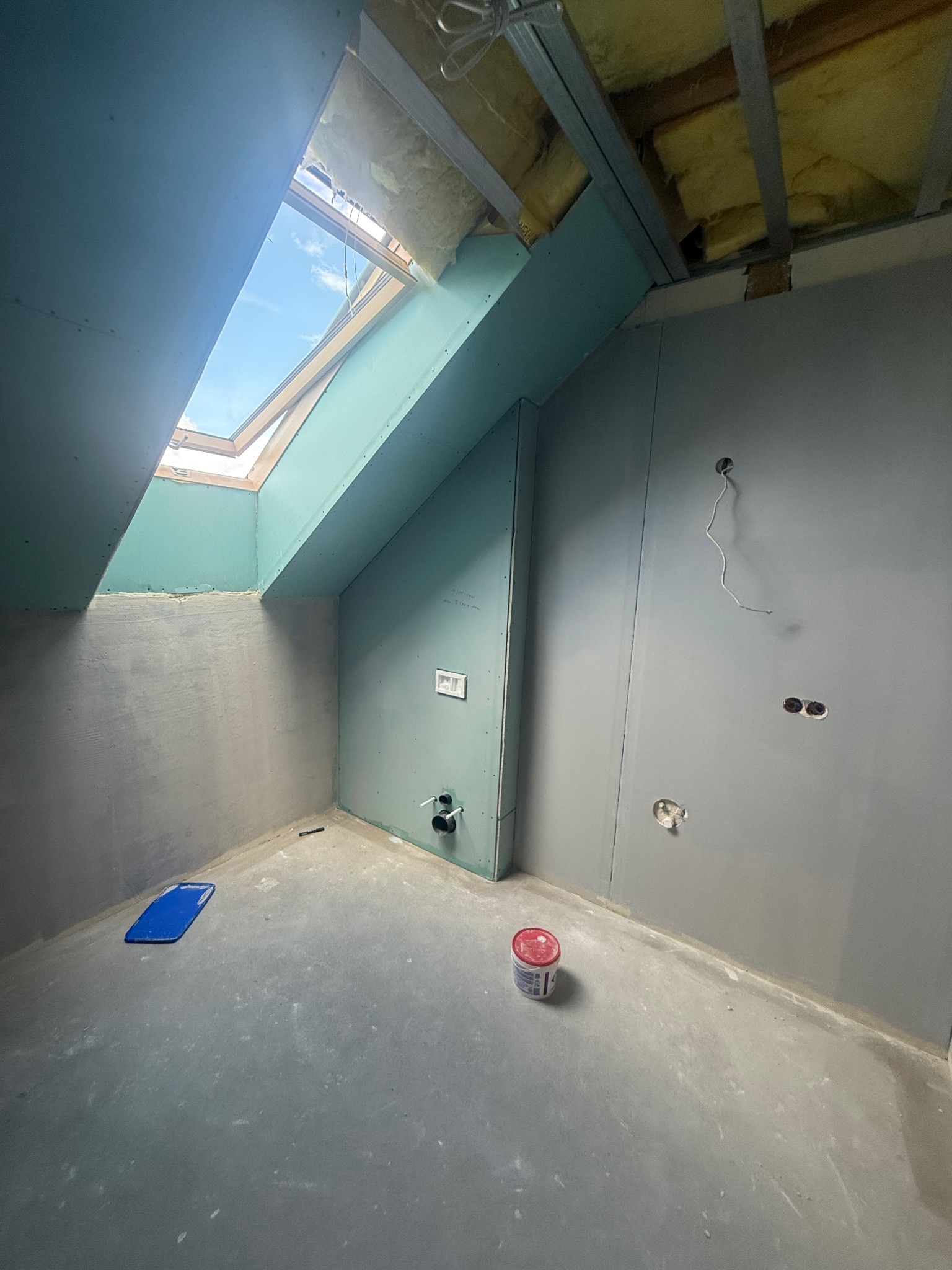 Poddasze w trakcie adaptacji: surowe ściany z płyt gips-karton, okno dachowe w skosie, widoczne instalacje elektryczne i hydrauliczne, przygotowane do dalszych prac wykończeniowych.