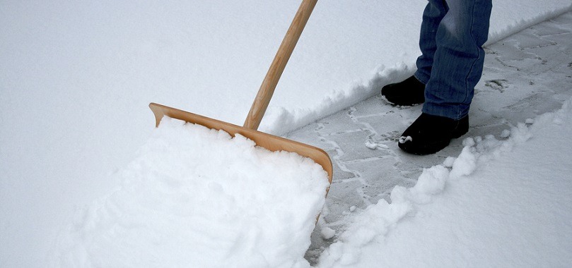 Osoba w jeansach odśnieża chodnik drewnianą łopatą. Widoczne czarne buty i fragment odśnieżonej kostki brukowej. Świeży śnieg.