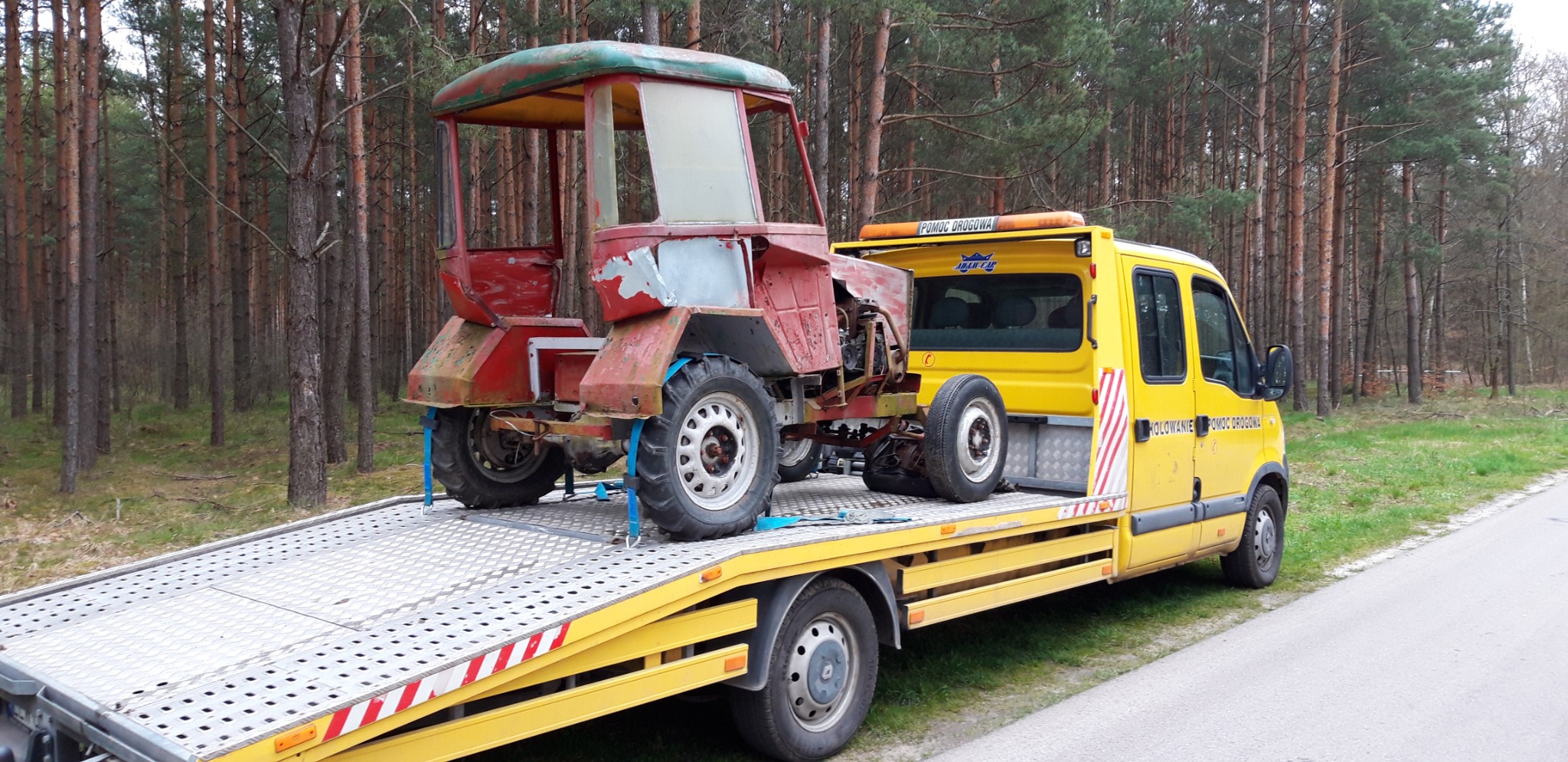 Żółta laweta z czerwonym, zardzewiałym traktorem na platformie, zabezpieczonym niebieskimi pasami, na tle leśnym. Widoczny napis 'Pomoc Drogowa'.