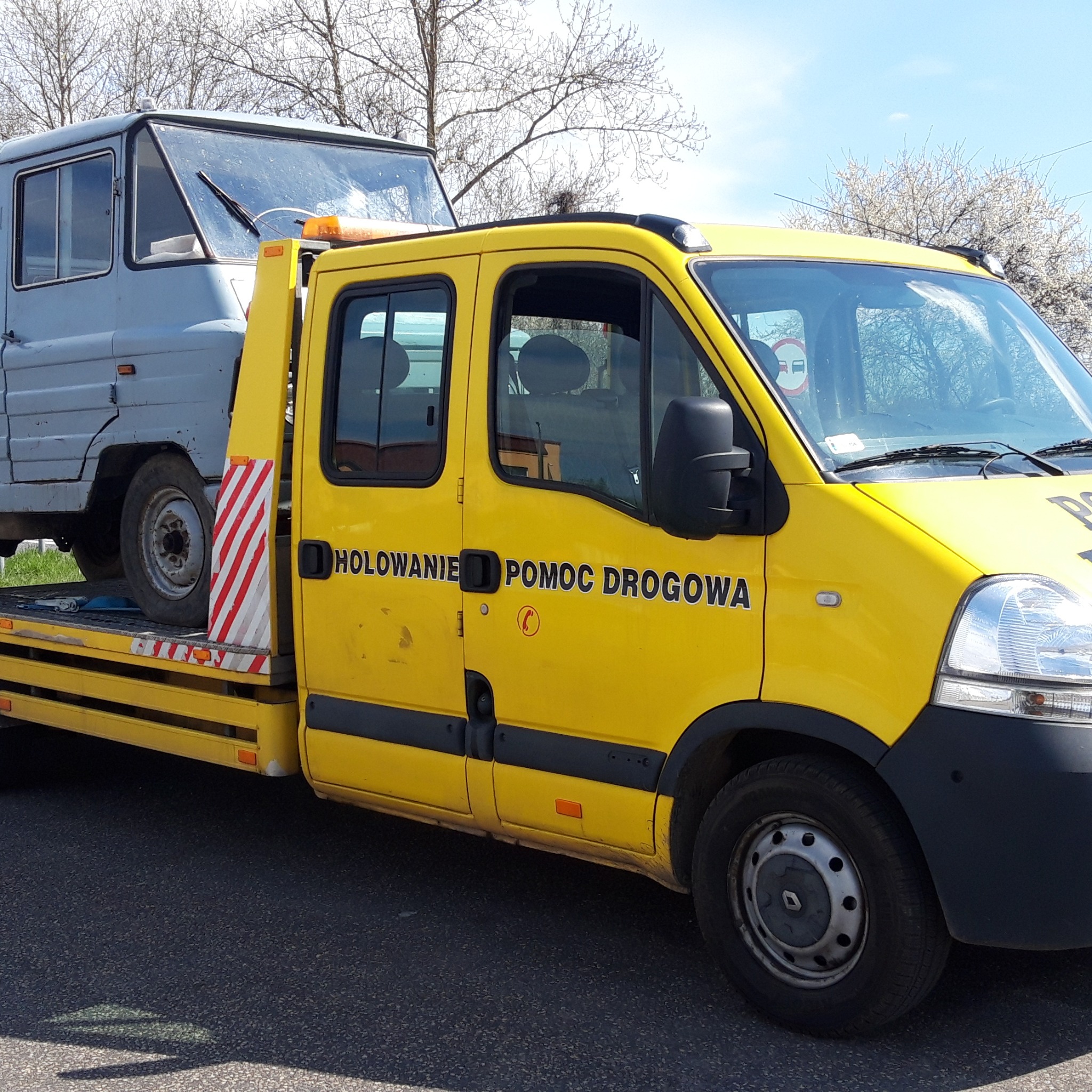 Żółta laweta z napisem 'Holowanie Pomoc Drogowa' przewozi szare auto na platformie. Widoczne drzewa w tle i fragment asfaltowej drogi.