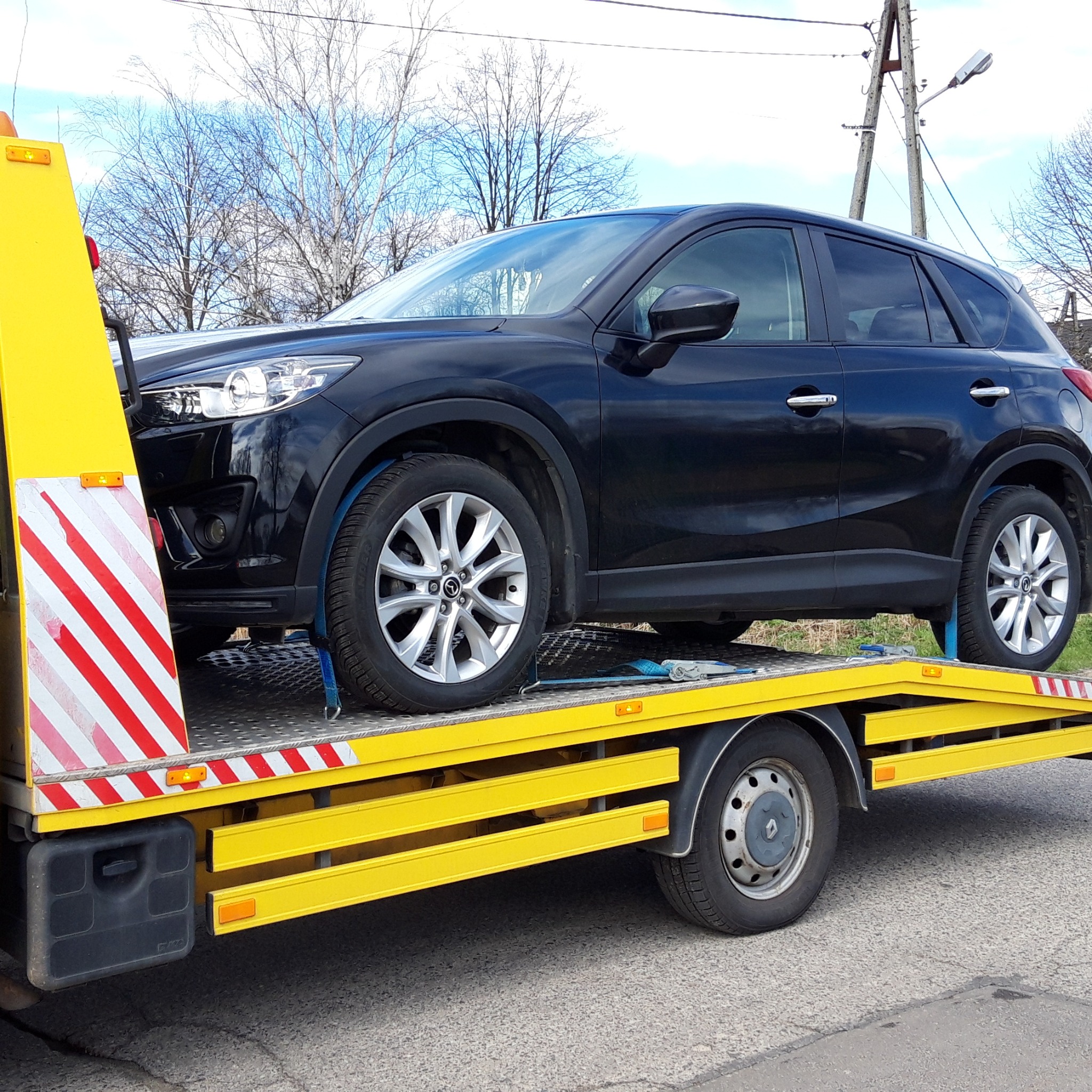 Czarny SUV na żółtej lawecie z czerwono-białymi pasami, zabezpieczony niebieskimi pasami transportowymi. Widoczne drzewa i słup energetyczny w tle.