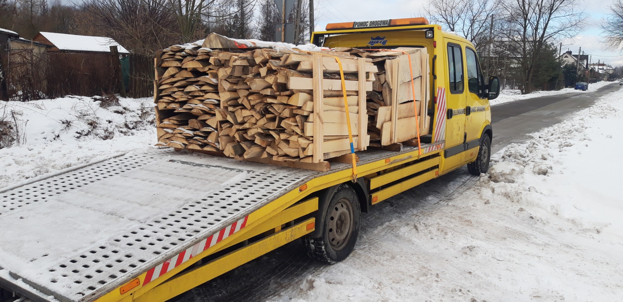 Żółta laweta z drewnem na platformie, zimowy krajobraz. Drewno zabezpieczone pasami. Widoczny napis 'Pomoc Drogowa' na pojeździe.