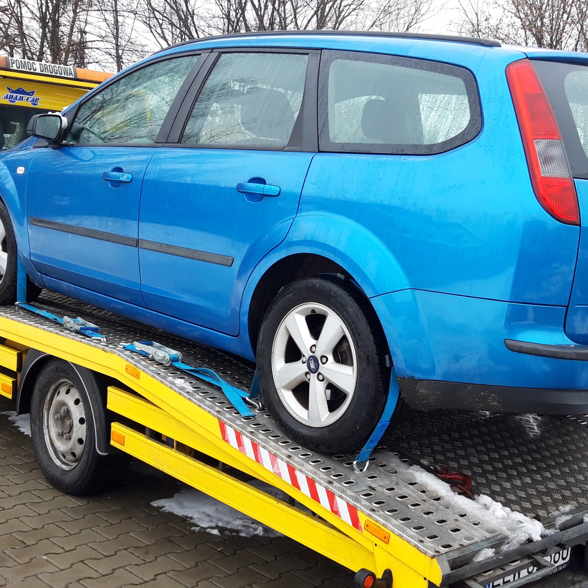 Niebieski samochód kombi zabezpieczony pasami na żółtej lawecie pomocy drogowej. Widoczne oznaczenia 'Pomoc Drogowa' i logo firmy na kabinie lawety.