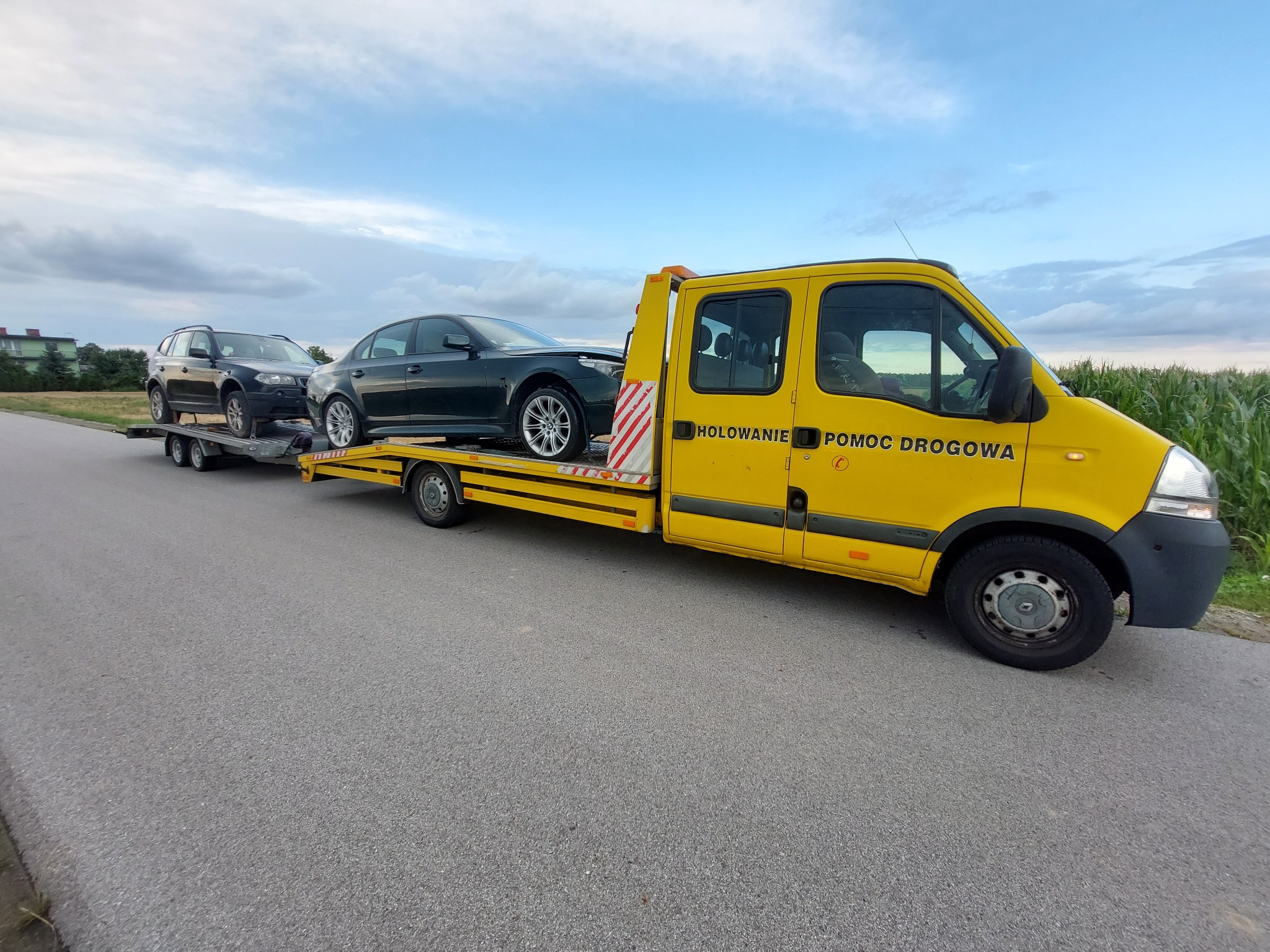 Żółta laweta z dwoma samochodami osobowymi na platformie, zaparkowana na asfalcie. Widoczny napis 'HOLOWANIE POMOC DROGOWA'.
