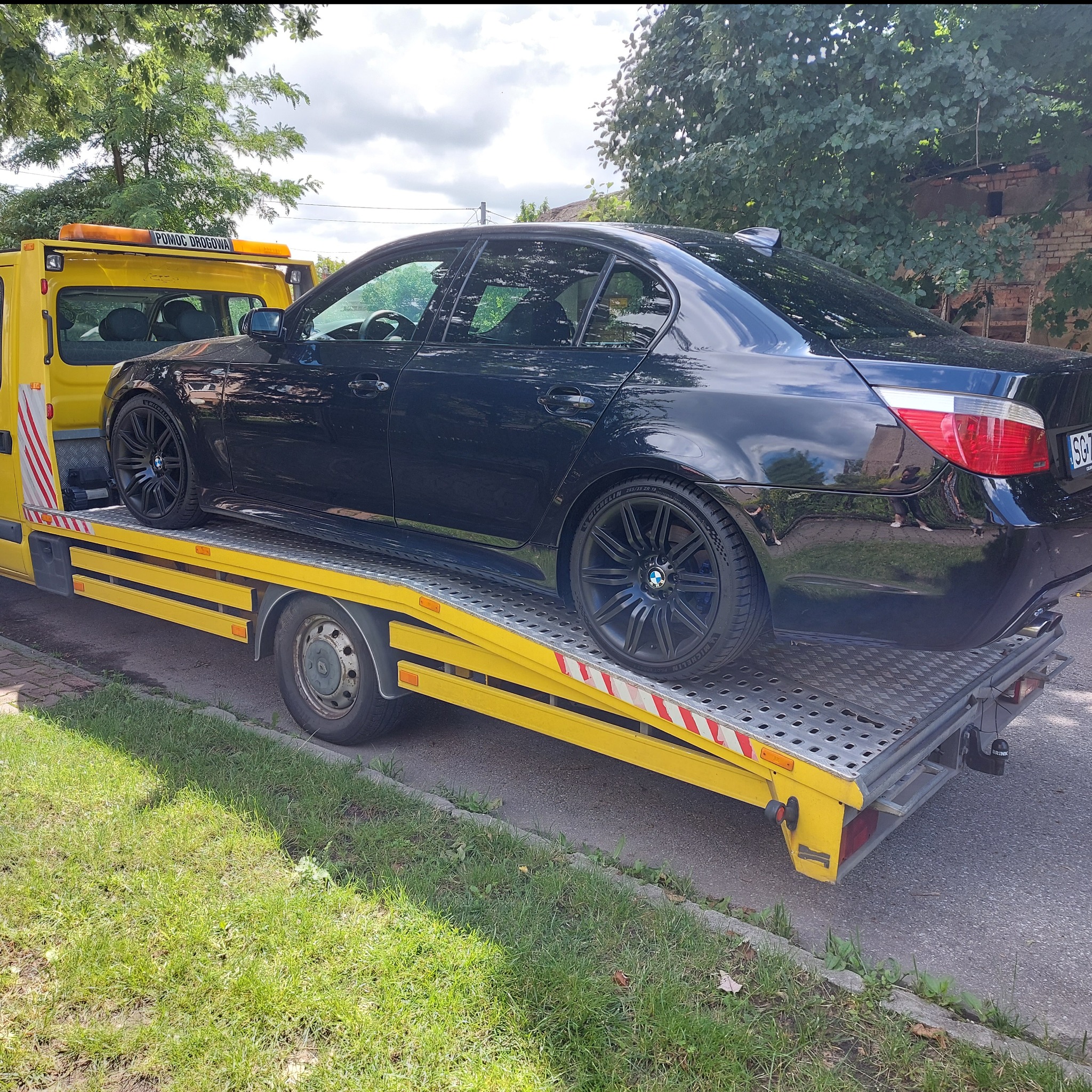 Czarny samochód BMW na żółtej lawecie pomocy drogowej, widok z boku, tablica rejestracyjna widoczna, drzewa w tle.