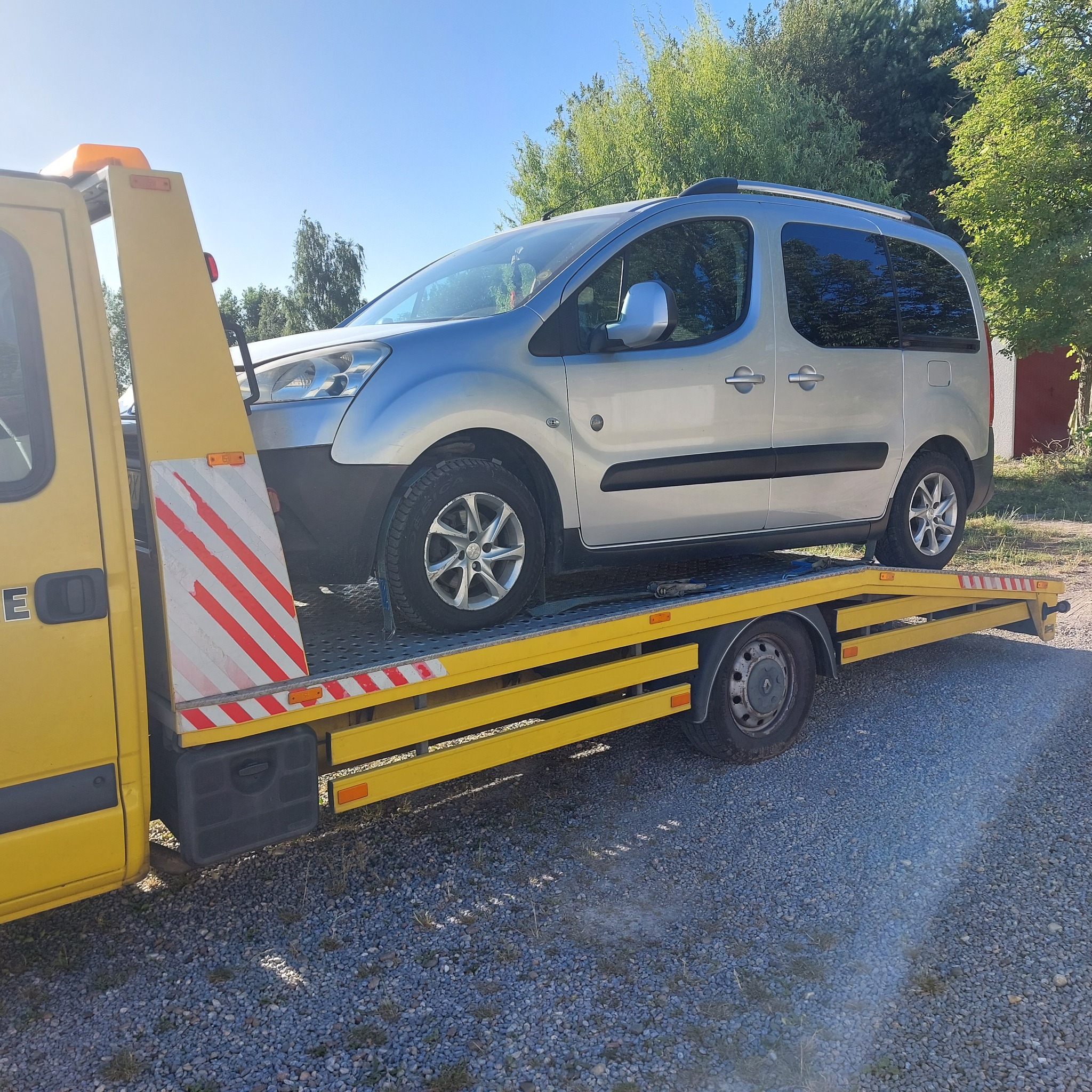 Srebrny samochód osobowy na żółtej lawecie z pomarańczowym światłem ostrzegawczym, widok z boku na tle zieleni i błękitnego nieba.
