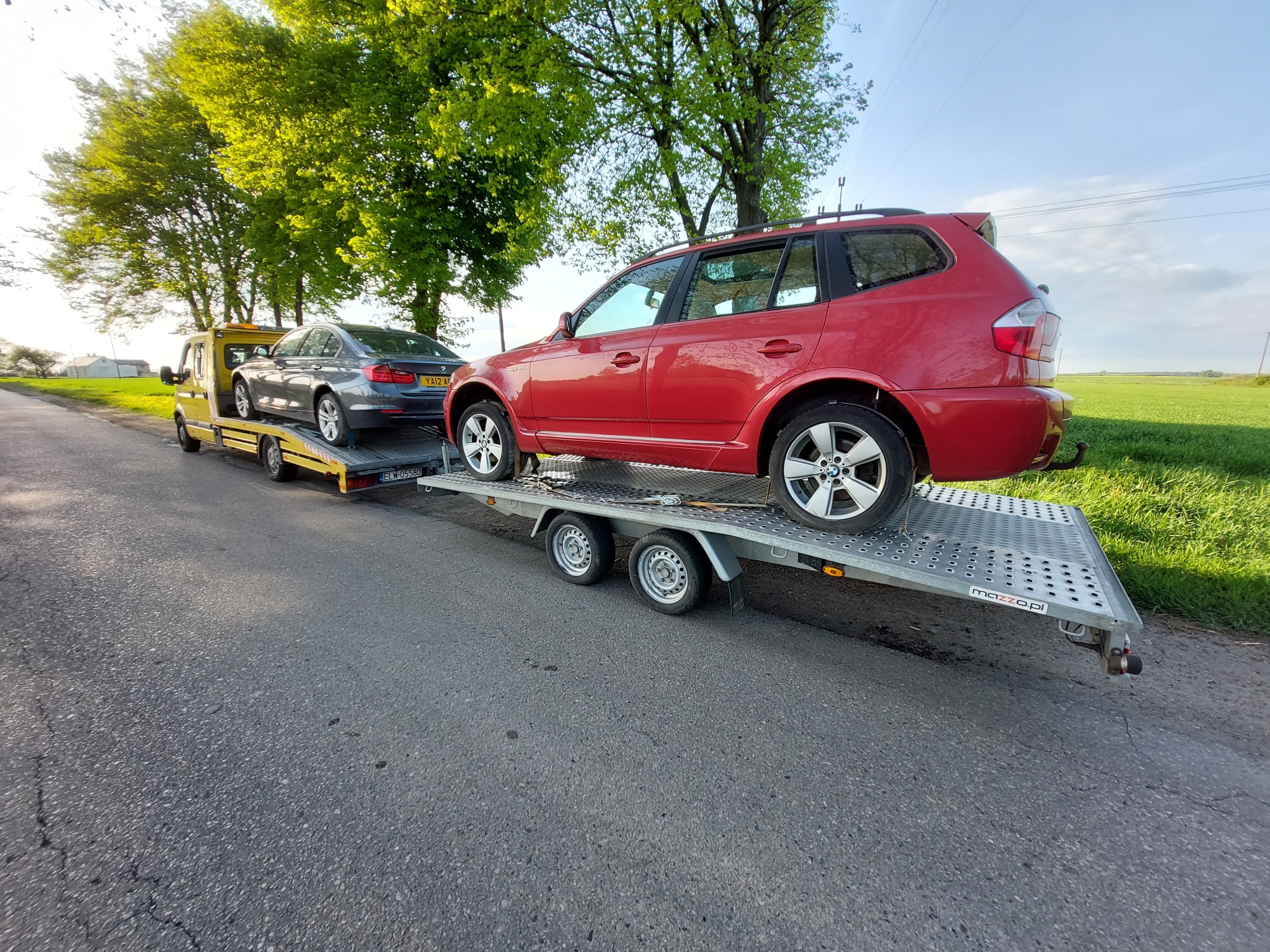 Żółta laweta z dwoma samochodami osobowymi na platformie, czerwony SUV na drugim planie, szary sedan na pierwszym. Transport pojazdów na drodze asfaltowej.