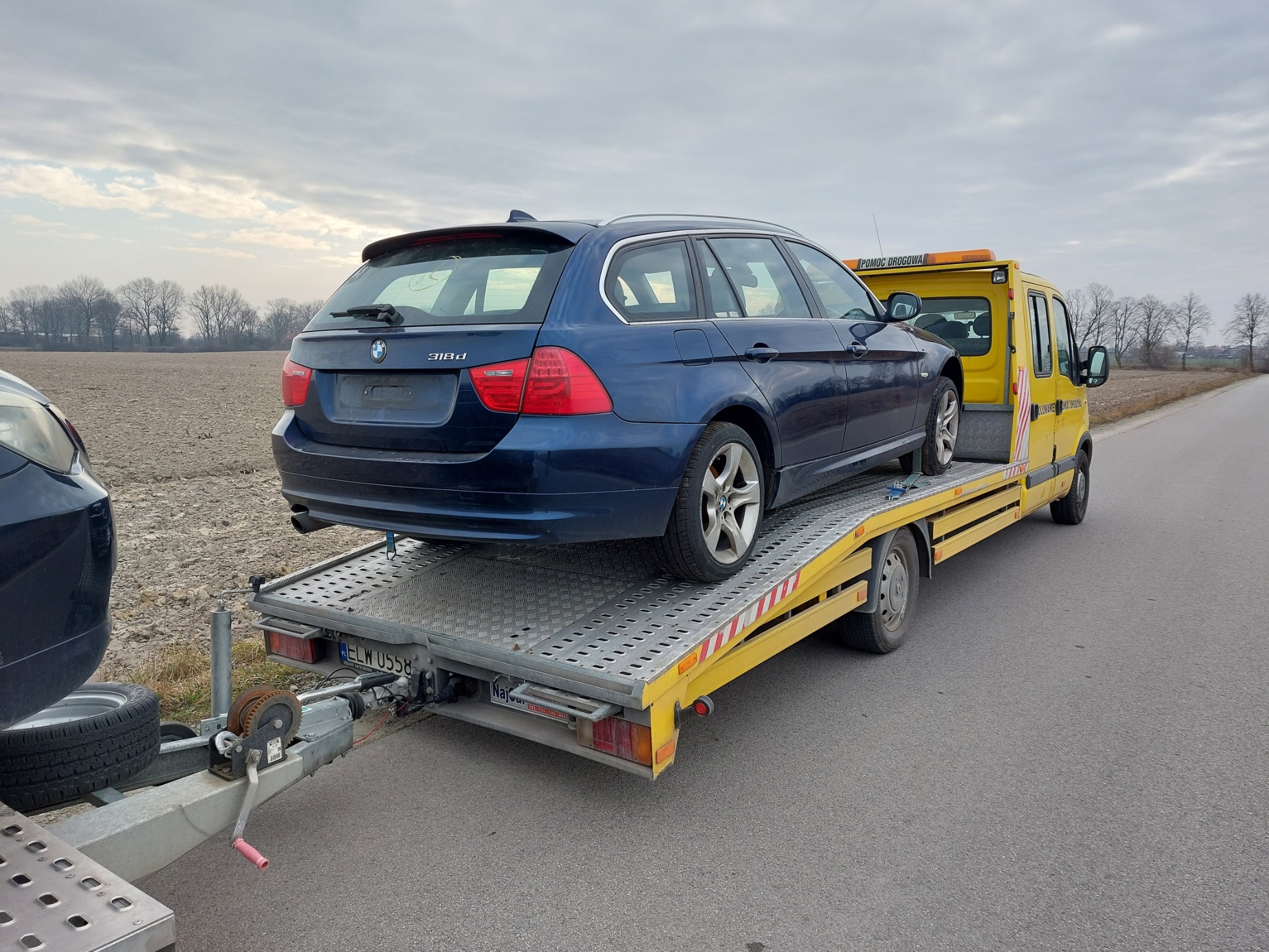 Niebieskie BMW 318d kombi na żółtej lawecie z napisem 'Pomoc Drogowa' na tle pola i pochmurnego nieba. Widoczny hak holowniczy i tablica rejestracyjna.