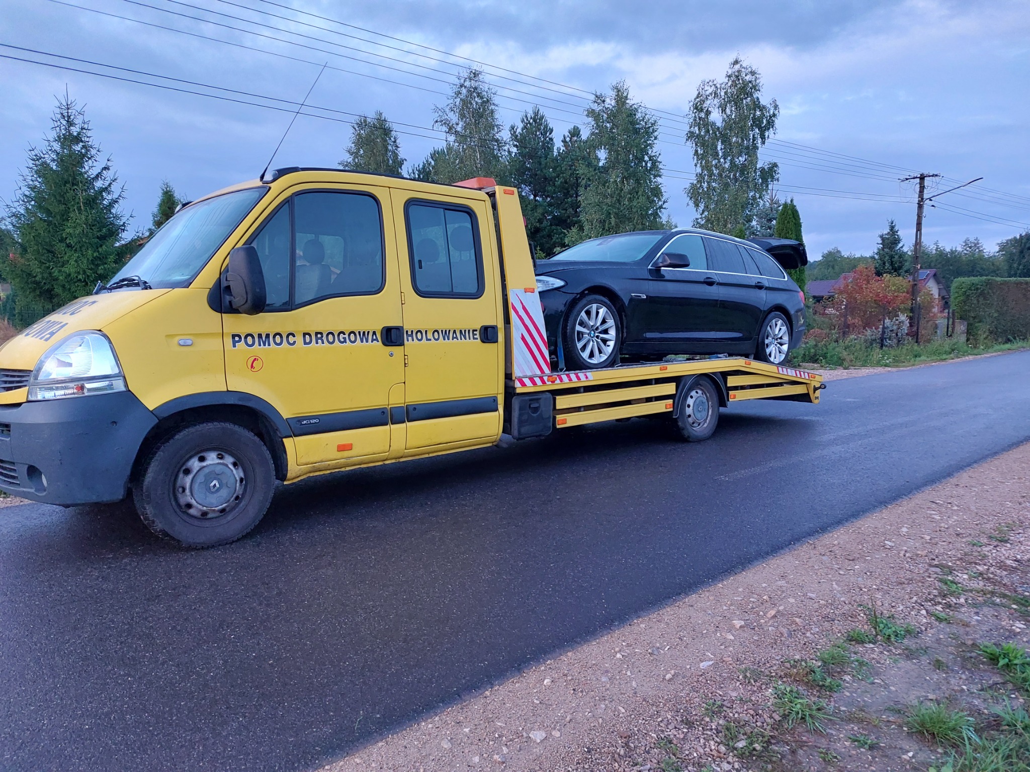 Żółta laweta z czarnym kombi BMW na platformie, zaparkowana na mokrej drodze. Widoczne napisy 'Pomoc Drogowa Holowanie'. Tło stanowią drzewa i domy.