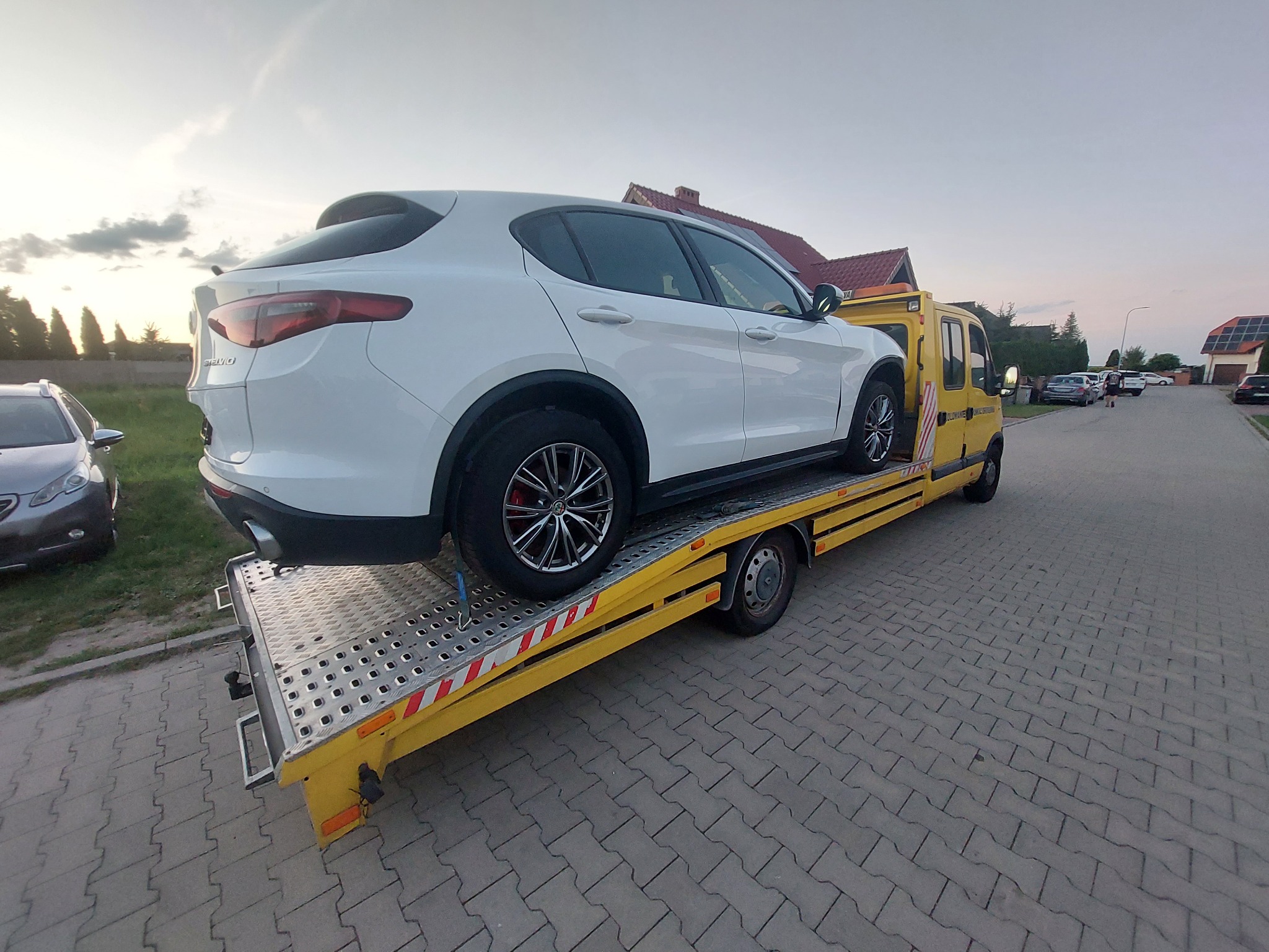 Żółta laweta z białym SUV-em Alfa Romeo Stelvio na platformie, zaparkowana na brukowanej drodze w spokojnej okolicy z domami w tle, widok z niskiej perspektywy.