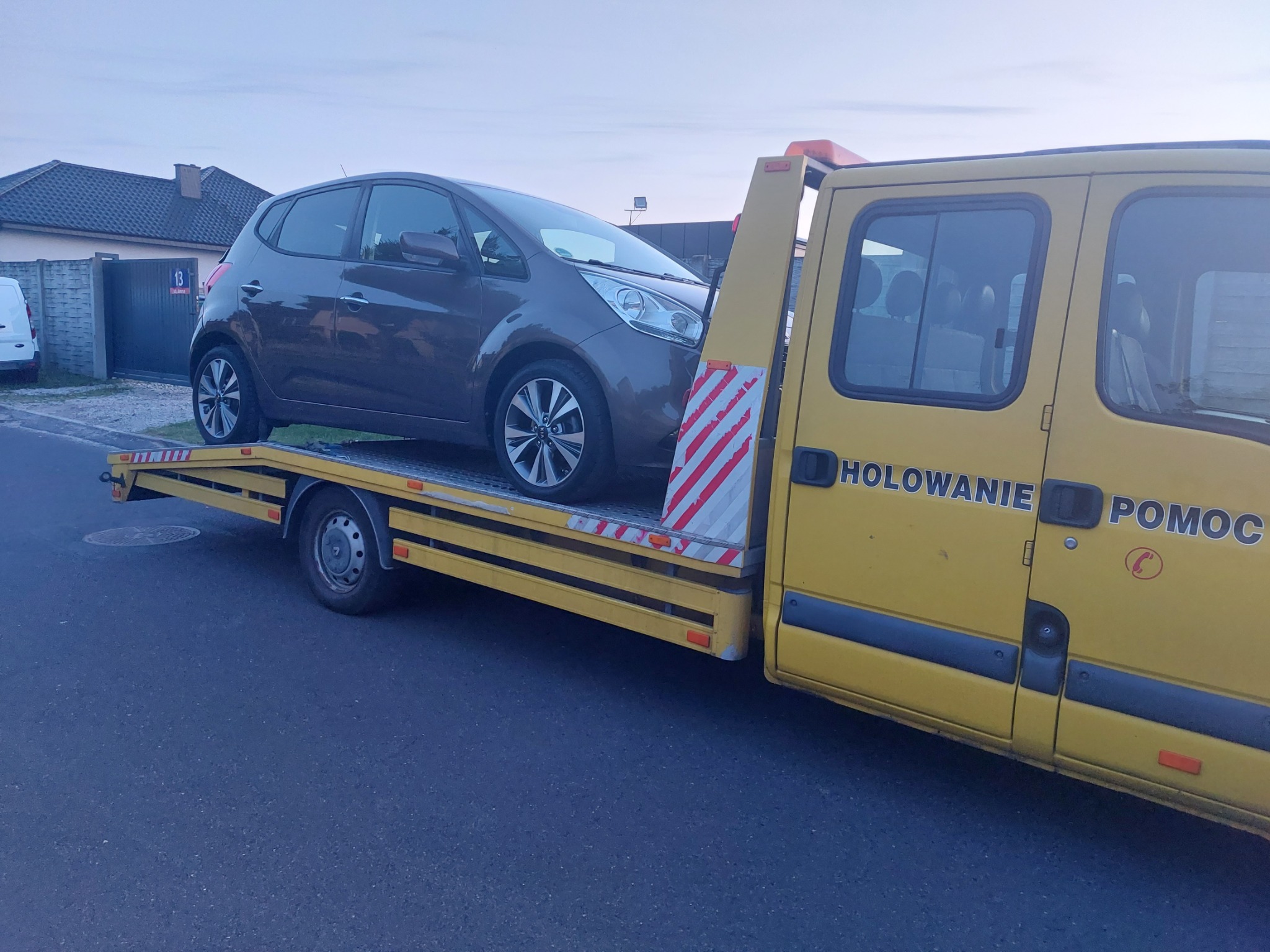 Brązowy samochód osobowy na żółtej lawecie z napisem 'HOLOWANIE POMOC' na tle ulicy i domu. Widok z boku, lekko z dołu.