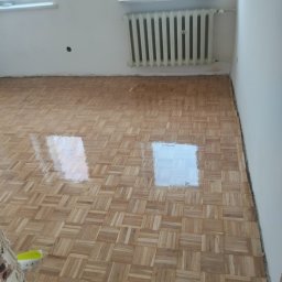Pogotowie parkieciarskie - Parkiet w jodełkę po lakierowaniu, z widocznymi refleksami światła. W tle grzejnik i okno, ściany w kolorze złamanej bieli. Fragment ściany w lewym dolnym rogu w trakcie wykończenia.