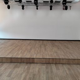 Pogotowie parkieciarskie - Sala z parkietem imitującym drewno, oświetleniem estradowym i białymi ścianami. Podwyższona scena z widocznym stopniem. Okno z żaluzjami po lewej stronie.