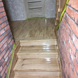 Pogotowie parkieciarskie - Drewniane schody po cyklinowaniu, zabezpieczone lakierem, z widoczną taśmą malarską przy ceglanej ścianie i parkiecie. Połyskująca powierzchnia.