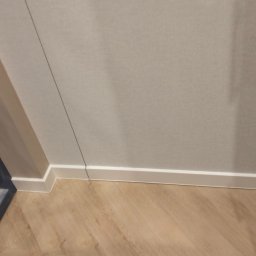 Pogotowie parkieciarskie - Jasne panele podłogowe ułożone w jodełkę, białe listwy przypodłogowe przy ścianie w odcieniach beżu, fragment ramy okiennej w lewym dolnym rogu.