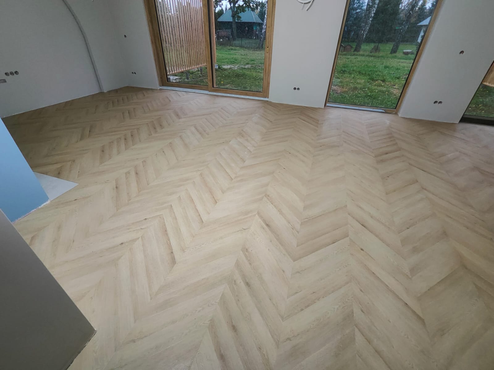 Jasny parkiet ułożony we wzór jodełki w przestronnym pokoju z dużymi oknami. Widoczne detale wykończenia ścian i naturalne światło wpadające przez okna.