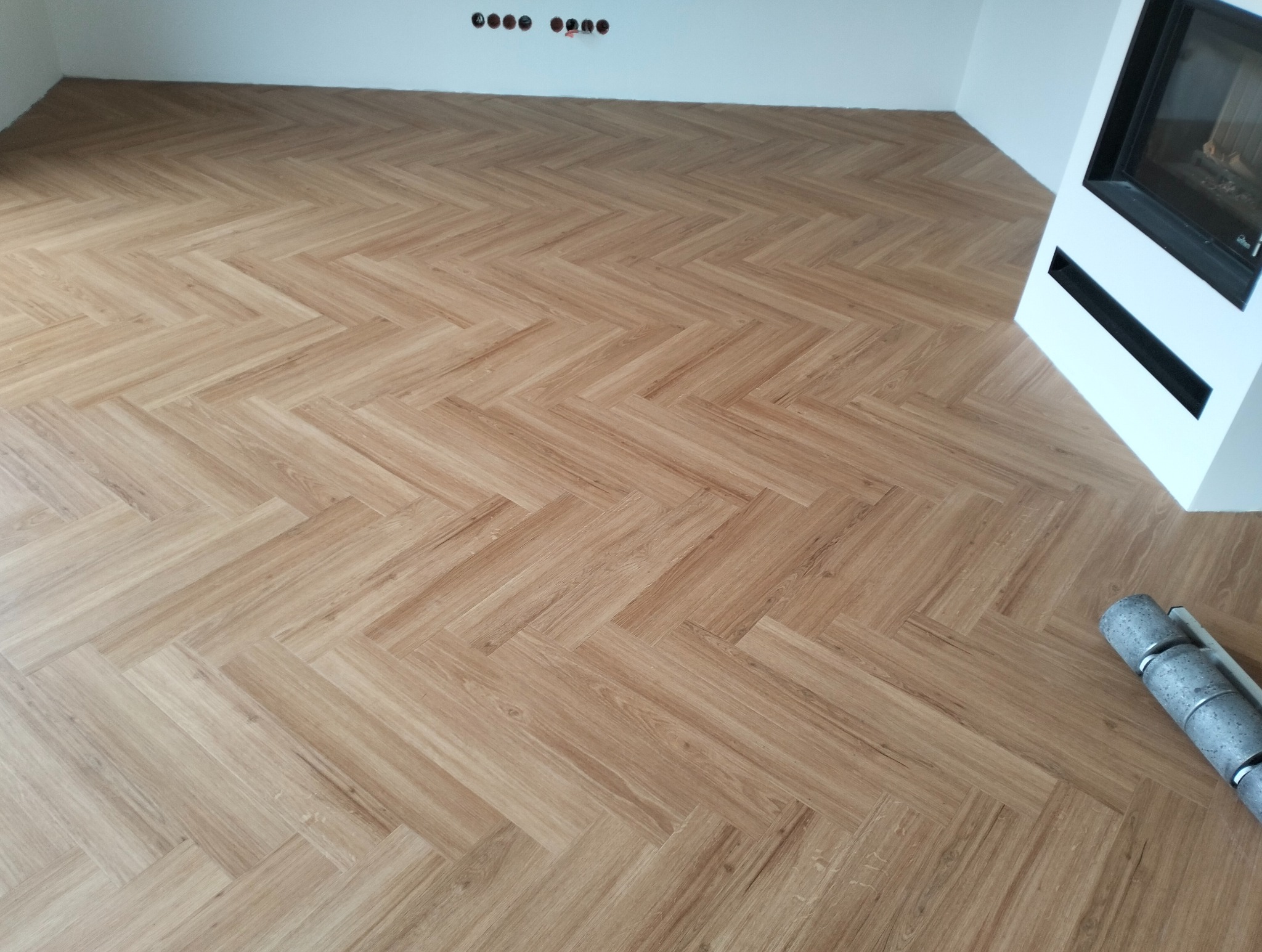 Jasny parkiet ułożony we wzór jodełki francuskiej w nowym salonie z kominkiem. Widoczne narzędzie do montażu paneli.