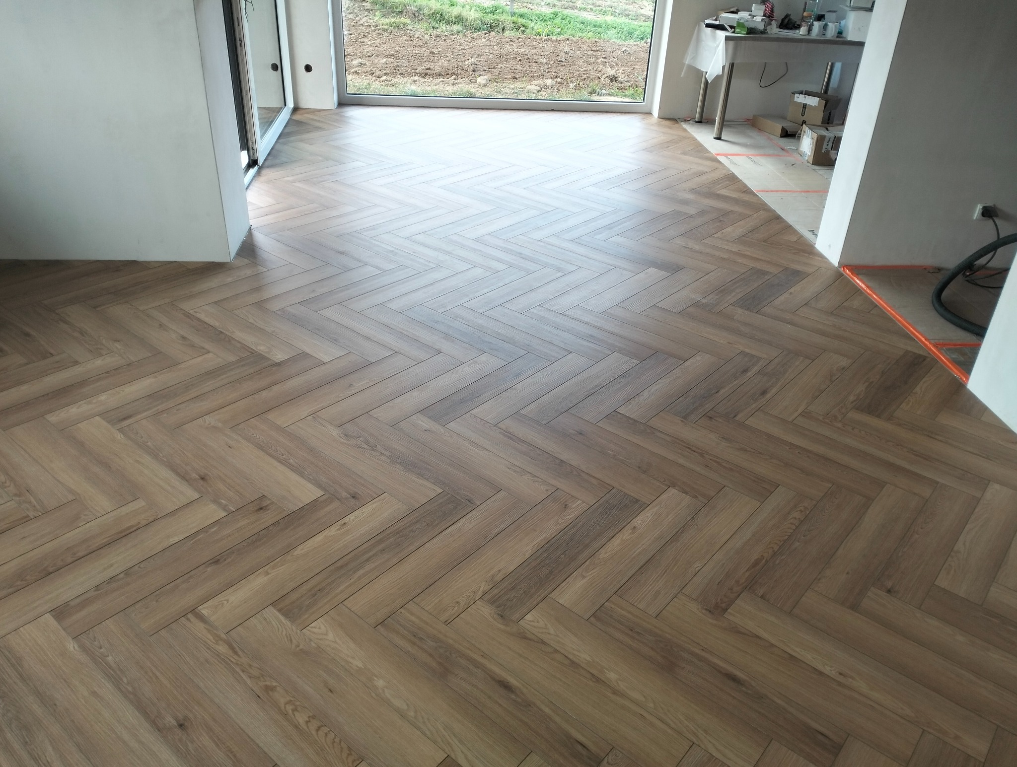 Ułożony parkiet w jodełkę francuską, jasne drewno, widok z góry, duże okno w tle. Częściowo widoczne przygotowania do dalszych prac wykończeniowych.