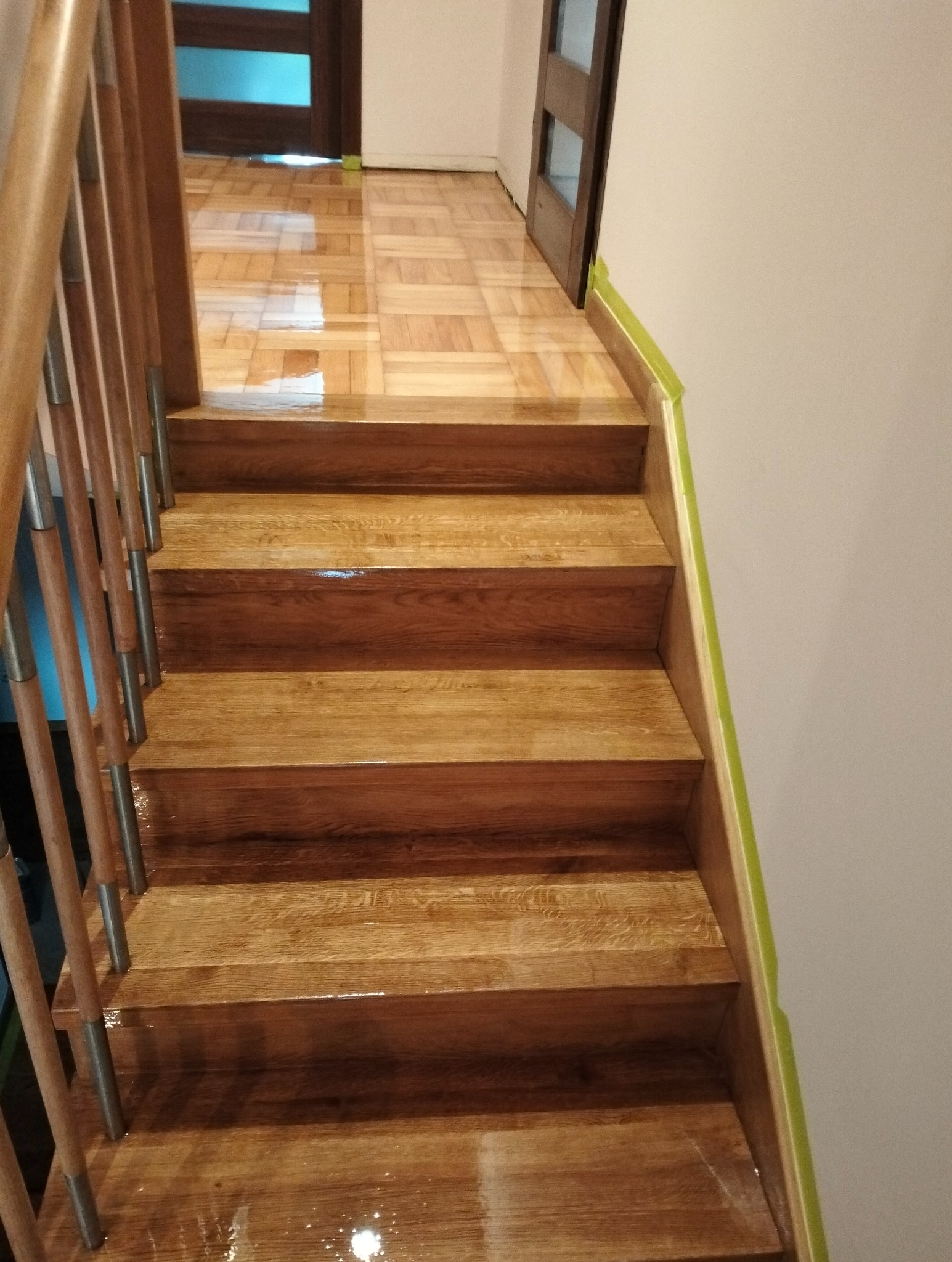 Błyszczące, drewniane schody po renowacji, z widoczną balustradą i fragmentem parkietu w tle. Świeżo nałożony lakier podkreśla naturalny rysunek drewna.