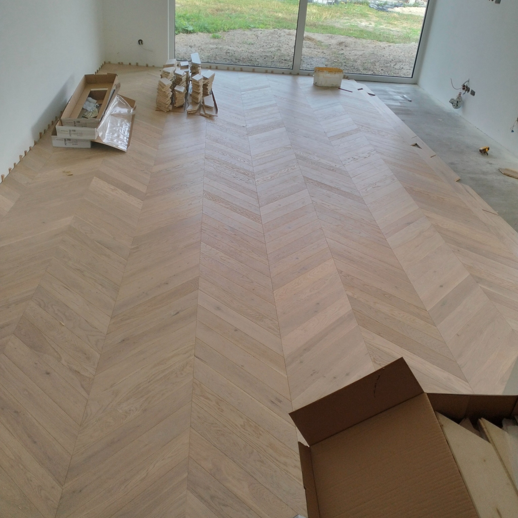 Jasny parkiet ułożony we wzór jodełki francuskiej w przestronnym wnętrzu. Widoczne paczki z panelami i listwy przypodłogowe. Duże okno z widokiem na zewnątrz.