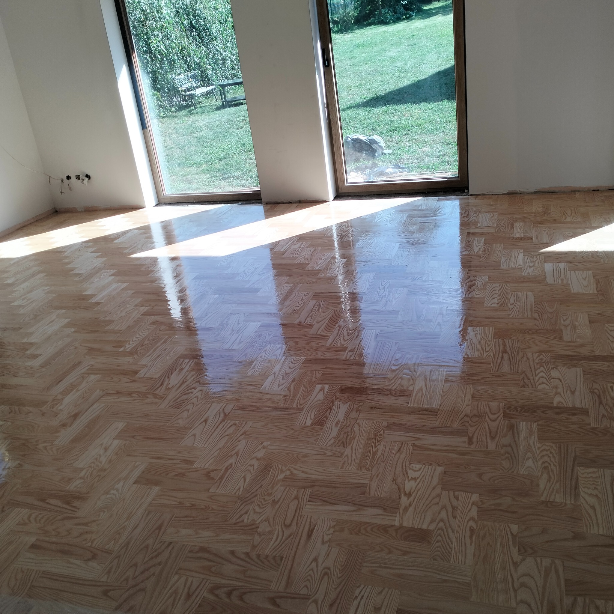 Ułożony parkiet w jodełkę, lakierowany na wysoki połysk, odbijający światło z okna. Widok na zielony ogród przez duże okno balkonowe.