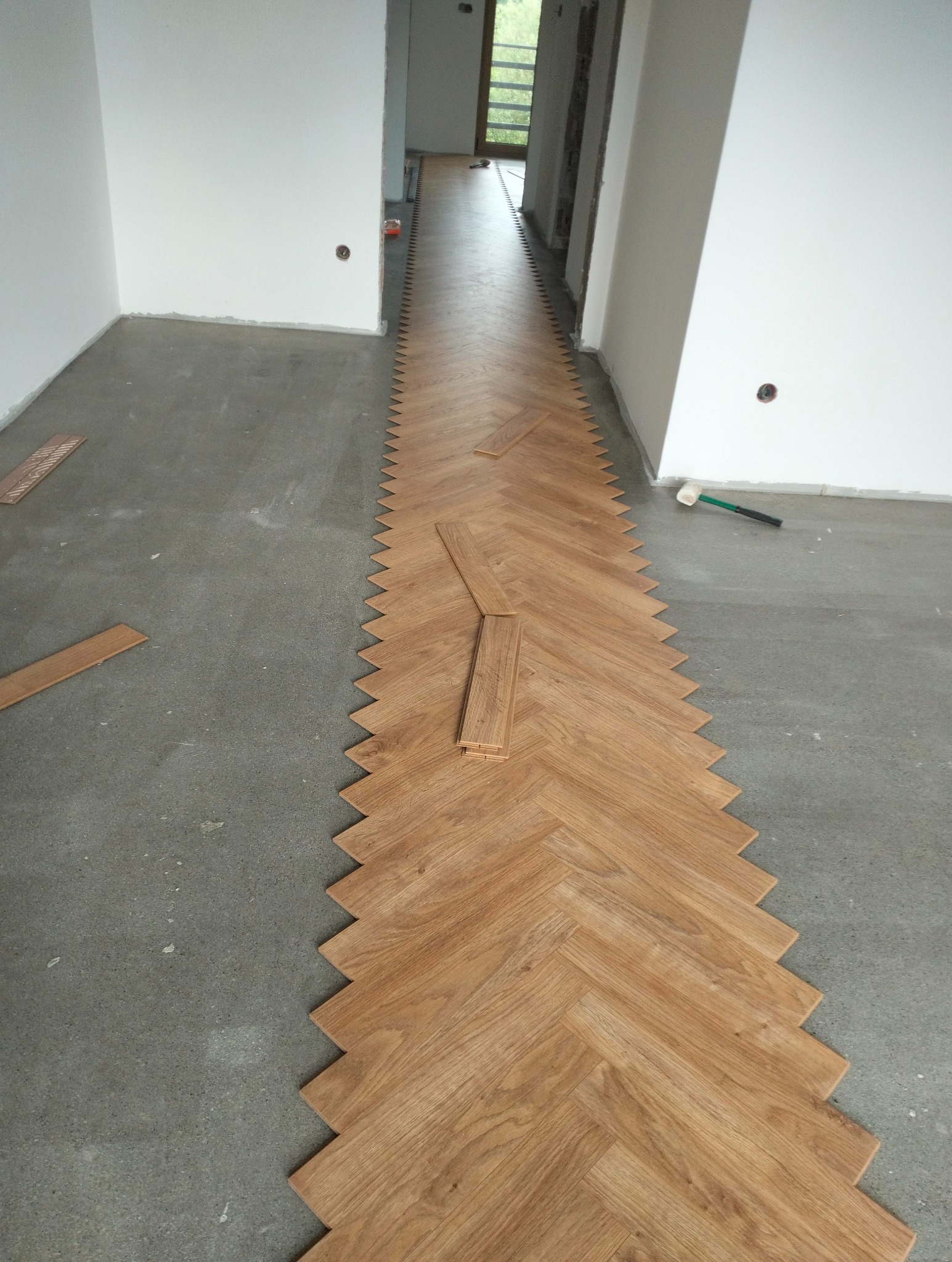 Wzorzyste układanie paneli podłogowych w jodełkę na surowym podłożu, widoczne narzędzia i niedokończone fragmenty. Jasne wnętrze w trakcie remontu.