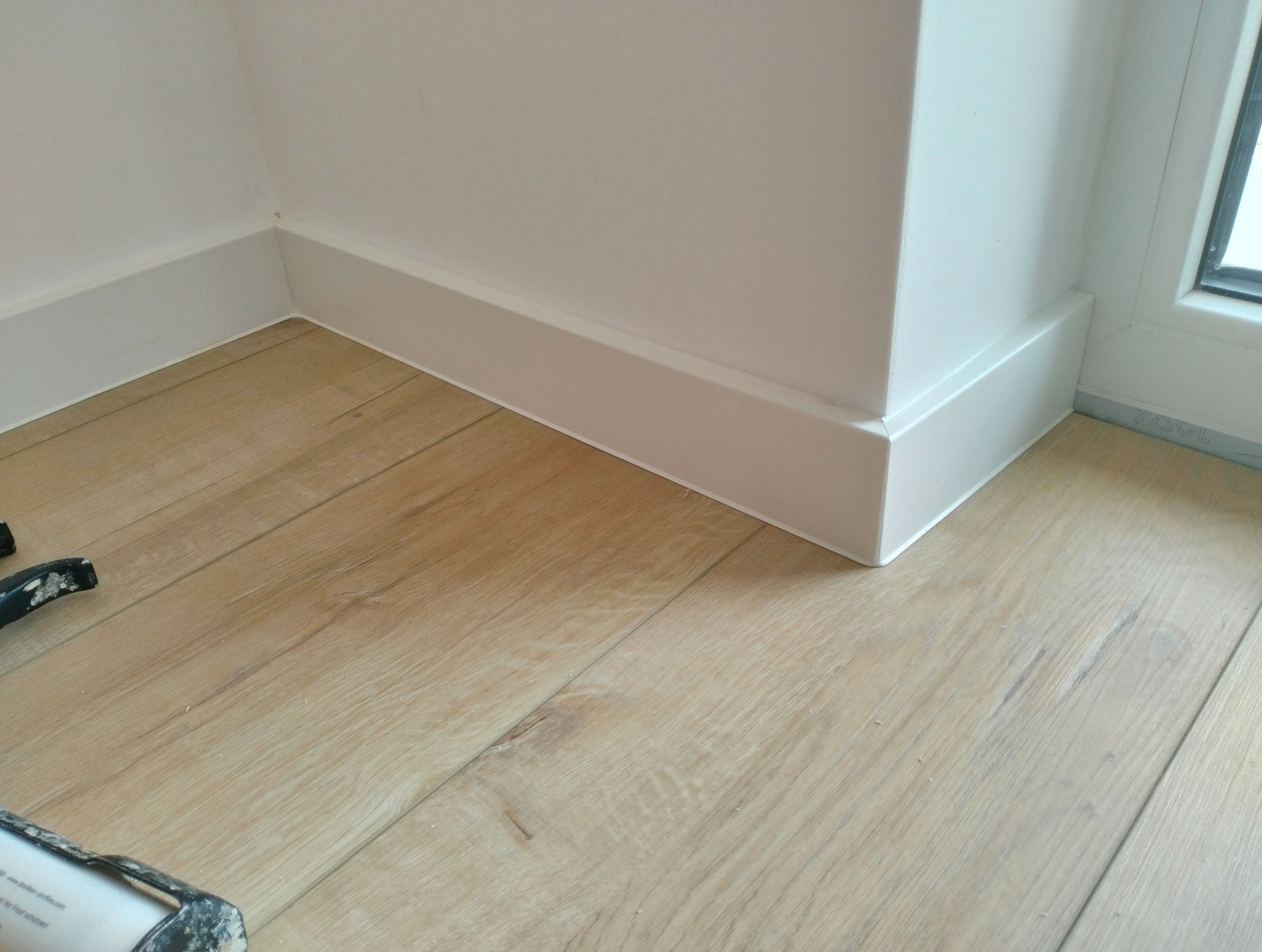 Jasne panele podłogowe z listwami przypodłogowymi w rogu pomieszczenia, widoczne narzędzie montażowe w lewym dolnym rogu. Minimalistyczne wykończenie wnętrza.