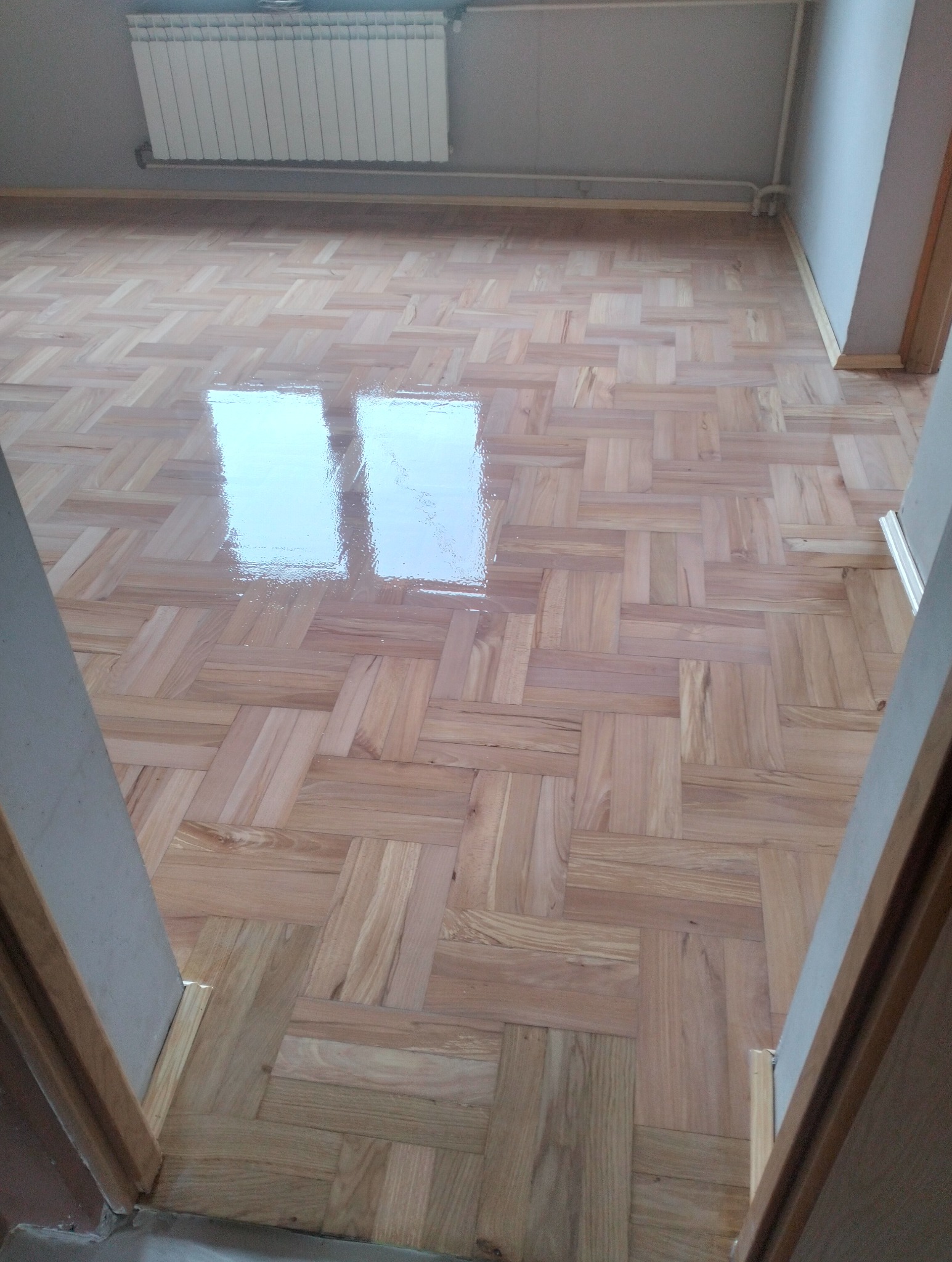 Parkiet ułożony w jodełkę, świeżo polakierowany, z widocznym odbiciem światła. Grzejnik na ścianie. Ujęcie z progu drzwi.