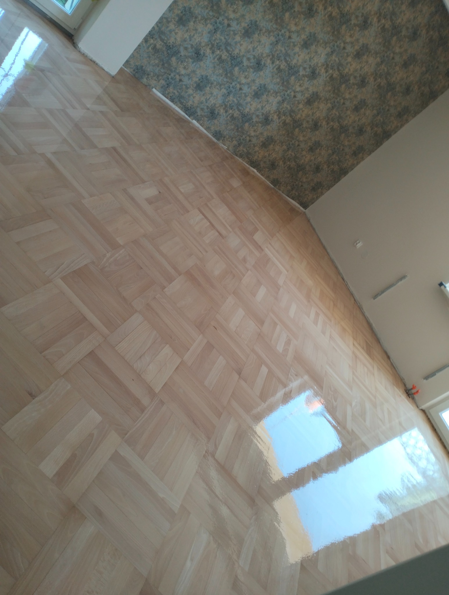 Ułożony parkiet w jodełkę, lakierowany na wysoki połysk, odbijający światło okien. Widoczny fragment ściany z tapetą w tle. Ujęcie z góry, ukośne.