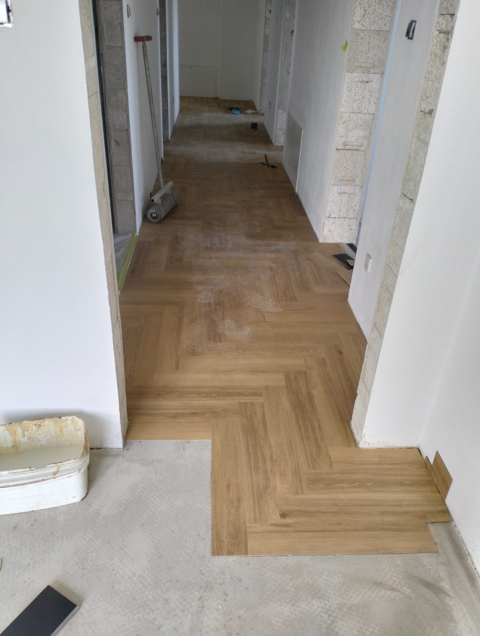 Układanie paneli w jodełkę w korytarzu. Widoczny fragment podłogi przed położeniem paneli, narzędzia i wiadro. Jasne wnętrze w trakcie remontu.