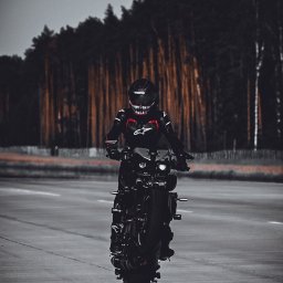 SKY Lens Radosław Nikonowicz - Mroczny motocyklista w czarnym kombinezonie i kasku z motywem zębów, jadący na czarnym motocyklu po asfalcie na tle lasu w ponurej kolorystyce.