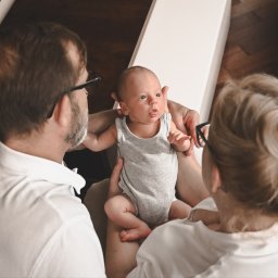 Sky Lens Fotografia - Rodzinne ujęcie z niemowlęciem w centrum, rodzice w białych koszulach patrzą na dziecko. Ciepłe światło, drewniane podłogi w tle. Urocza sesja rodzinna.