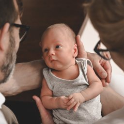 Sky Lens Fotografia - Rodzinne ujęcie: Noworodek w objęciach rodziców, z delikatnym uśmiechem i skupionym wzrokiem, uchwycone w ciepłym, naturalnym oświetleniu.