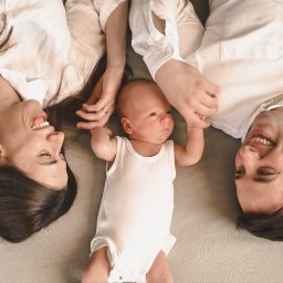 SKY Lens Radosław Nikonowicz - Uśmiechnięta rodzina leżąca na plecach: mama z rozpuszczonymi włosami, tata i niemowlę w białym body trzymające się za ręce. Zdjęcie z góry, ciepłe światło.